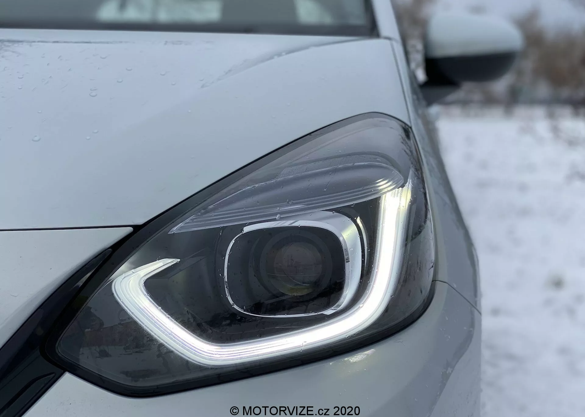 Detailný záber na predný ľavý svetlomet Honda Jazz 2020, ktorý ukazuje moderný dizajn s LED alebo HID osvetlením. Svetlomet obsahuje charakteristické denné svetlo v tvare háčika, projektorovú šošovku a kvapky vody naznačujúce nedávne zrážky alebo umývanie. Elegantný dizajn je zdôraznený hladkými líniami a kontúrami, s chladným alebo zimným pozadím, pravdepodobne so snehom.