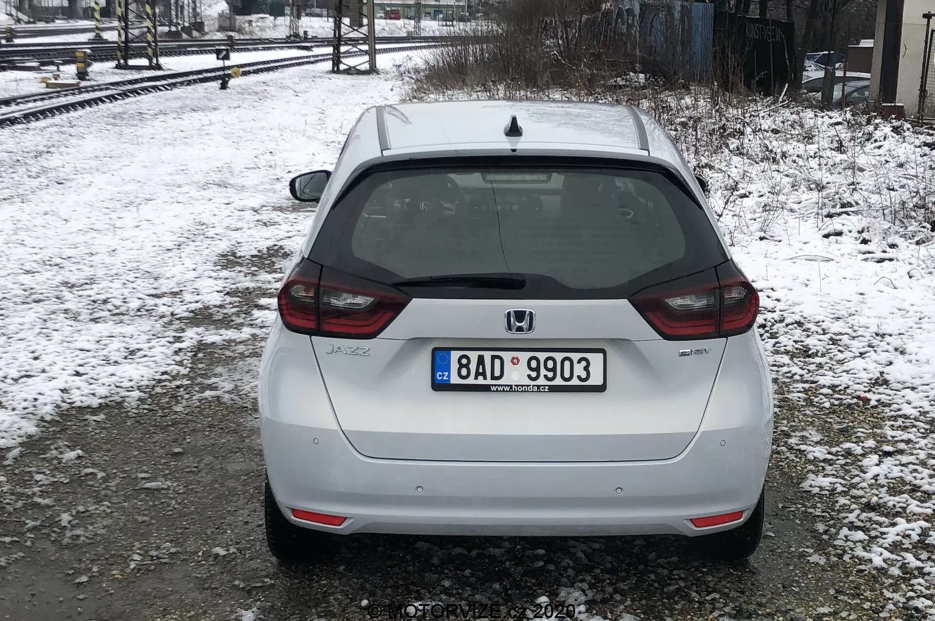Na obrázku je zadný pohľad na biely Honda Jazz 2020, zaparkovaný na zasneženom povrchu blízko železničných tratí v rozptýlených alebo zamračených svetelných podmienkach. Auto zaberá väčšinu rámu so stredovým zadným pohľadom, ktorý zobrazuje európsku poznávaciu značku, zadný stierač, vysoko osadené brzdové svetlo a znak Honda. Modelový odznak 'JAZZ' je viditeľný v dolnej ľavej časti kufra a nad poznávacou značkou sa nachádza zadná kamera. Zadné svetlá čiastočne obopínajú boky, s odrazovými svetlami na spodnom nárazníku, ktorý má tiež integrovanú čiernu časť. Stopy nečistôt naznačujú, že auto jazdilo po mokrých alebo blatistých cestách. Sneh a holé stromy naznačujú, že je zima alebo skorá jar.