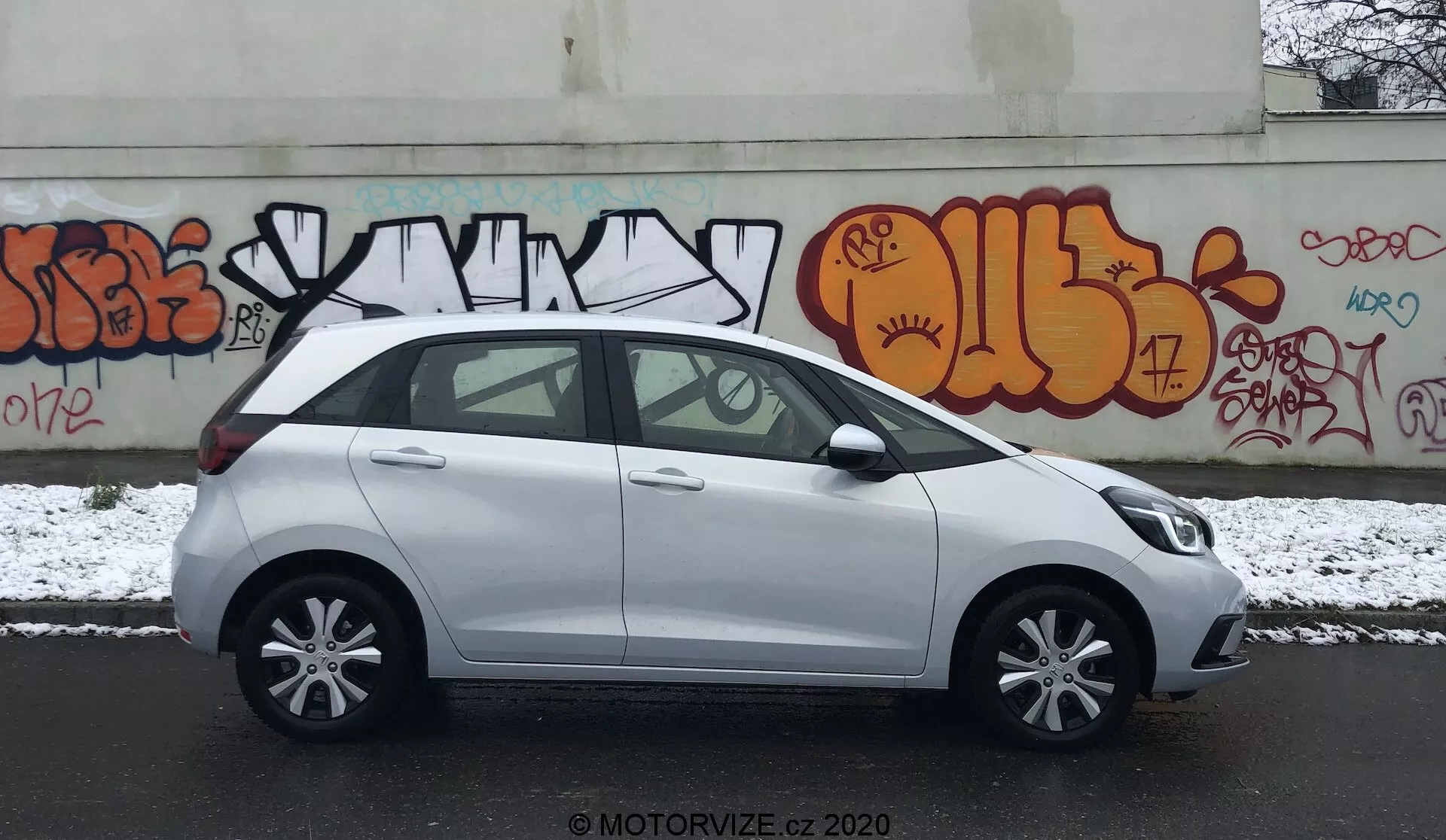 Bočný profil bieleho kompaktného hatchbacku Honda Jazz 2020 zaparkovaného na mestskej ulici s graffiti na pozadí. Scéna je počas zamračeného dňa s ľahkým popraškom snehu, čo naznačuje zimné podmienky. Obrázok zachytáva stranu vodiča s farebne zladenými kľučkami dverí, bočnými zrkadlami, päťlúčovými zliatinovými ráfikmi a štandardnými oknami s čiernym B-stĺpikom. Vozidlo vyzerá čisto s drobnými mokrými škvrnami od snehu. Viditeľný je vodoznak 'MOTORVIZE.CZ 2020', ktorý naznačuje zdroj fotografie.