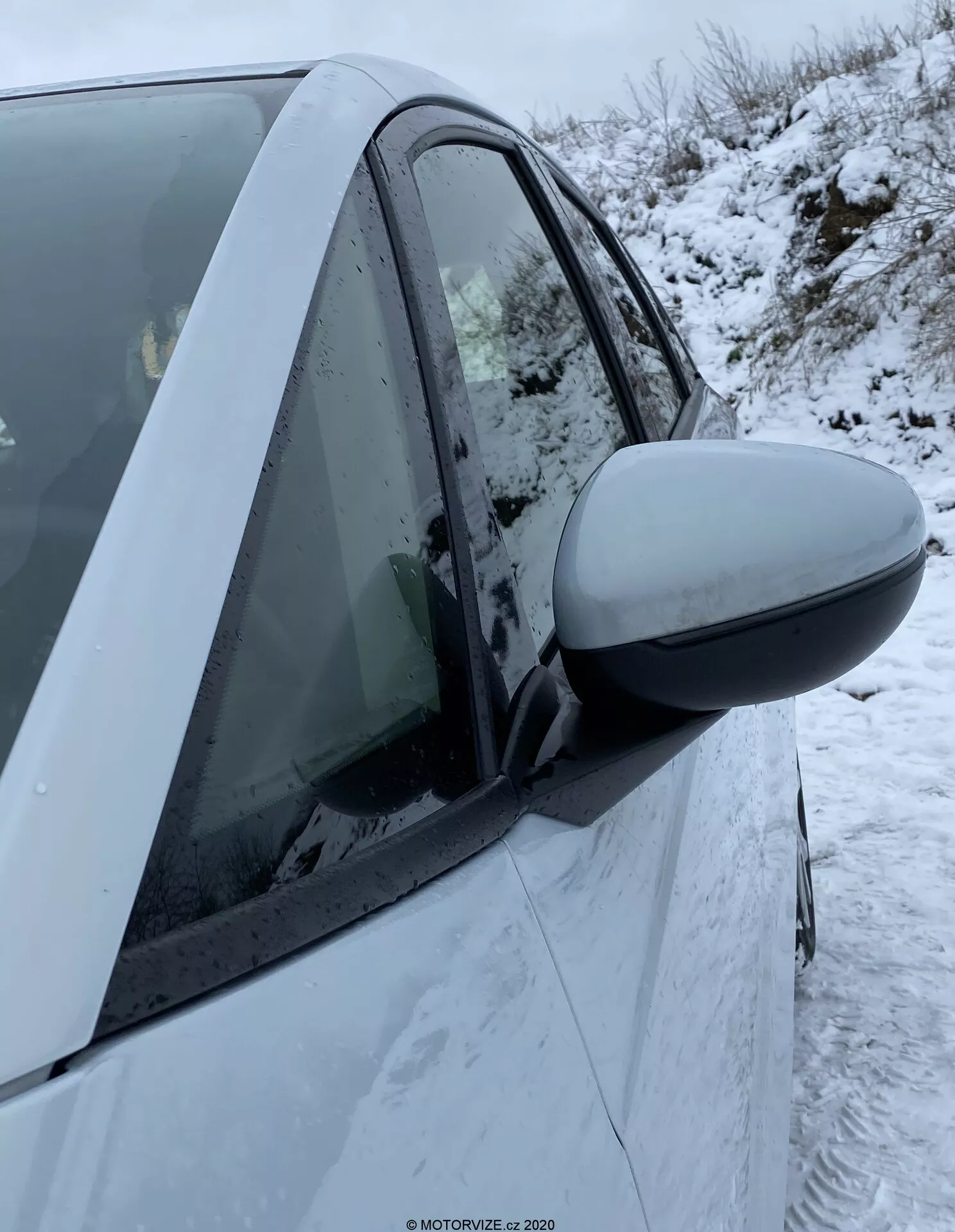Detailný záber na pravú stranu Honda Jazz 2020, zameraný na oblasť dverí vodiča. Nápadné bočné zrkadlo sa tiahne od ľavej strany obrázka do stredu, čo naznačuje nedávne alebo nadchádzajúce použitie. Kryt zrkadla zodpovedá farbe auta, čo naznačuje, že ide o štandardnú funkciu. Moderné dizajnové prvky zahŕňajú aerodynamické zrkadlo a neporušenú karosériu. Okno je vytiahnuté so zahmlením vo vnútri a na dverách a okne sú viditeľné snehové vločky. Viditeľný B-stĺpik a časti obidvoch dverí, predných a zadných, naznačujú štvor-dverové vozidlo. Bočný indikátor nie je viditeľný. Pozadie ukazuje zimné podmienky s viditeľným snehom na zemi.