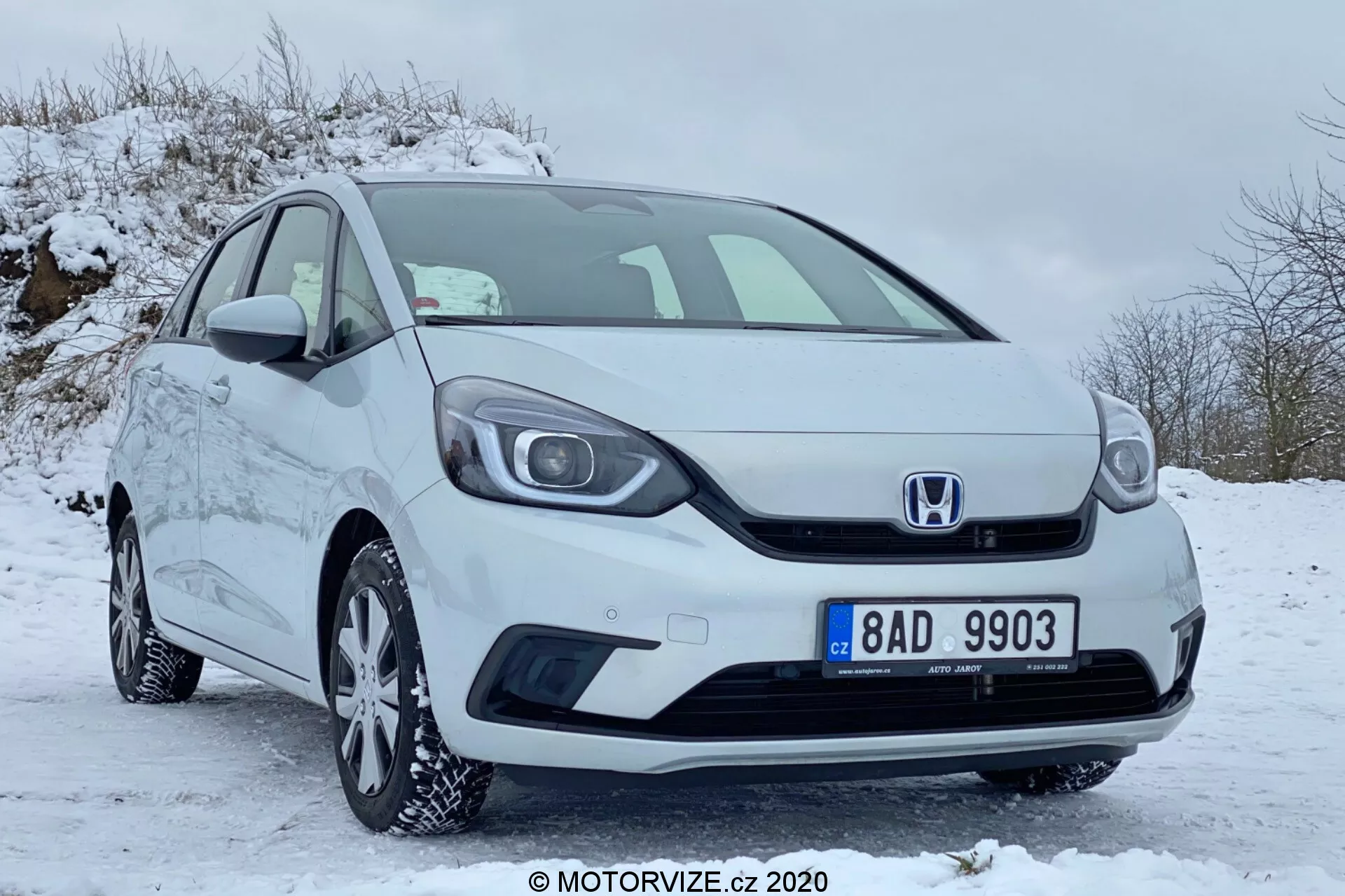 Predný trojštvrťový pohľad na biele kompaktné auto Honda Jazz 2020 zaparkované na zasneženej ploche, zobrazujúci mriežku s emblémom Honda, predný nárazník, svetlomety, zliatinové disky s zimnými pneumatikami, bočné zrkadlá a kľučky dverí, všetko v dobre udržiavanom a čistom stave.