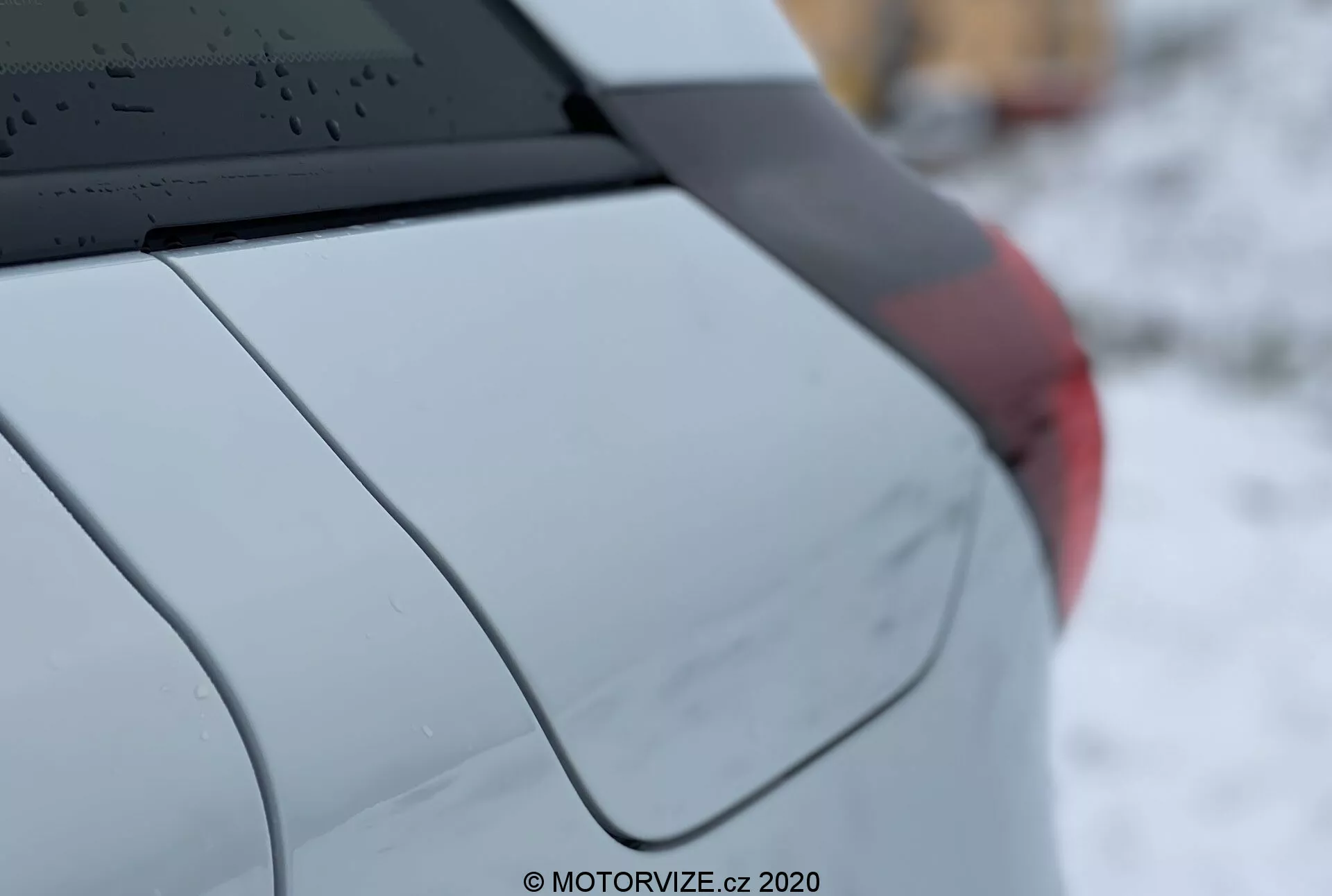 Detailný záber na Honda Jazz 2020, ukazujúci pravý zadný štvrtý auta z troch štvrtinového pohľadu zboku. Vlastnosti zahŕňajú zadné okno s odmrazovacími čiarami, anténu v tvare plutvy žraloka namontovanú na streche, C-stĺpik zdôrazňujúci dizajn auta, hladko zakrivený zadný blatník, fragment zadného svetelného zostavy a metalický alebo perleťový lak. Malá hĺbka ostrosti zdôrazňuje detaily vozidla, zatiaľ čo pozadie je rozmazané. Vodotlač číta 'MOTORVIZE.cz, 2020.'