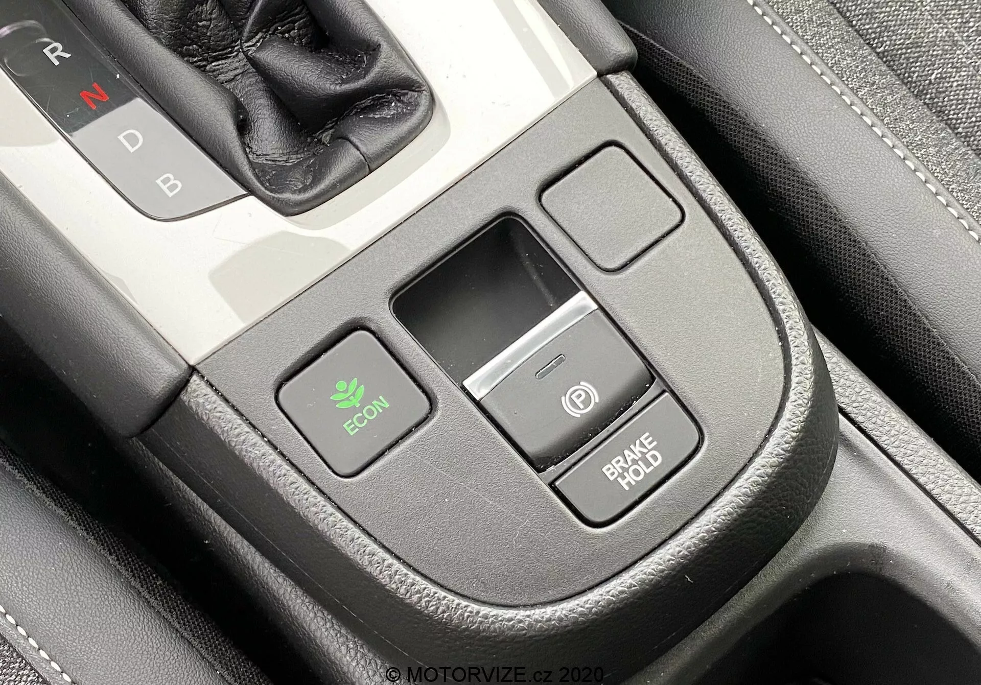 Detailný záber stredovej konzoly interiéru vozidla Honda Jazz 2020 z mierne vrchného uhla z pohľadu vodiča. Selektor prevodovky s 'N' pre Neutrál je čiastočne viditeľný vpravo hore, čo naznačuje automatickú prevodovku. Na ľavej strane je tlačidlo ECON pre ekonomický režim, v strede malý obdĺžnikový úložný priestor a na pravej strane sú dve stohované tlačidlá—jedno s ikonou napájania a druhé označené 'BRAKE HOLD', ktoré bráni vozidlu v pohybe pri zastavení. Konzola má prémiový povrch s koženým vzhľadom a ozdobným prešívaním.