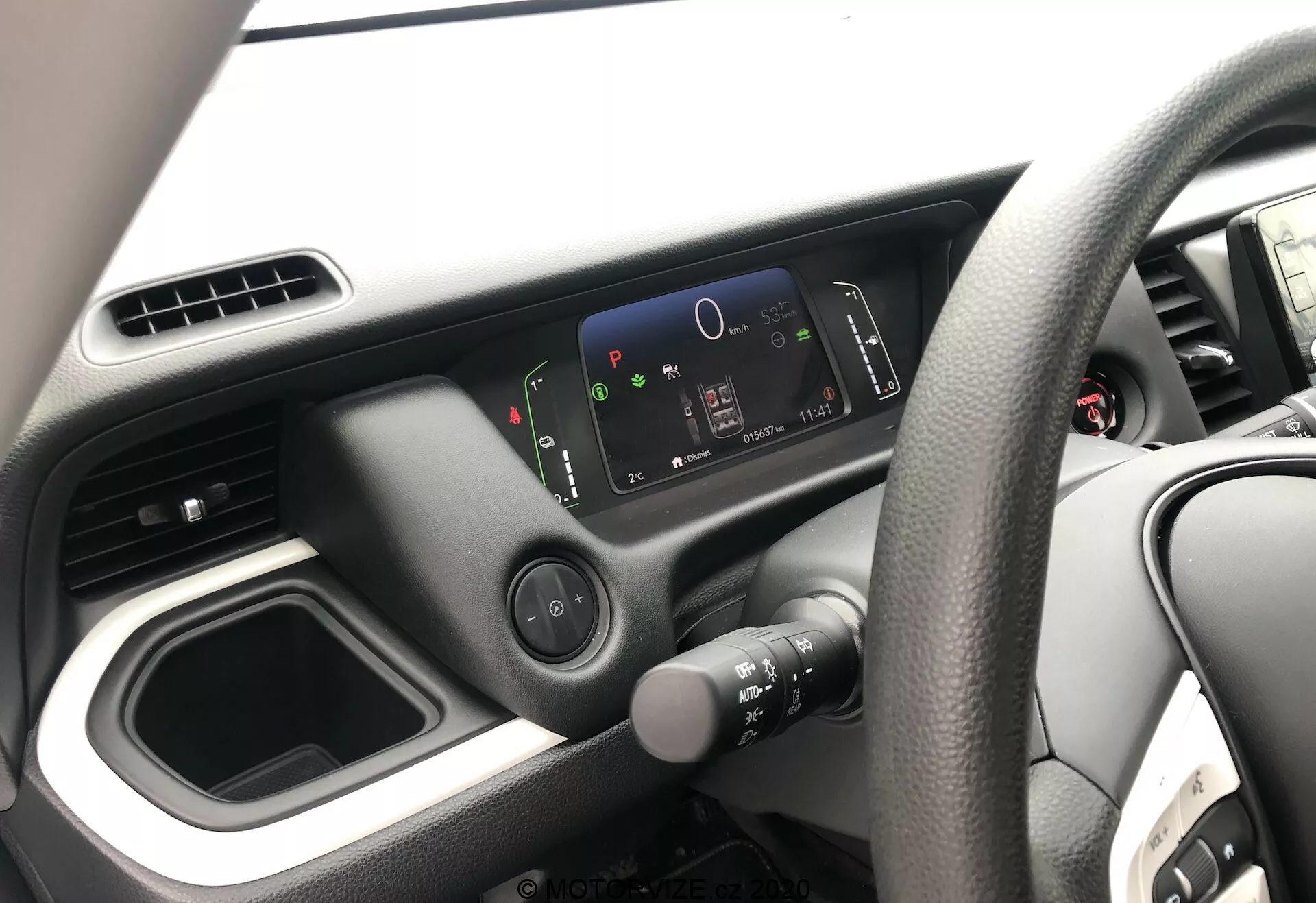 Pohľad na interiér modelu Honda Jazz 2020, zameraný na stranu vodiča z pohľadu pasažiera. Obrázok zobrazuje volant na pravej strane s dizajnom so štyrmi ramenami, minimalistickú prístrojovú dosku s digitálnym displejom s vysokým rozlíšením a jednoduché rozloženie stredovej konzoly s prázdnym úložným priestorom a ventilačnými otvormi. Viditeľná je iba časť dverí vodiča.