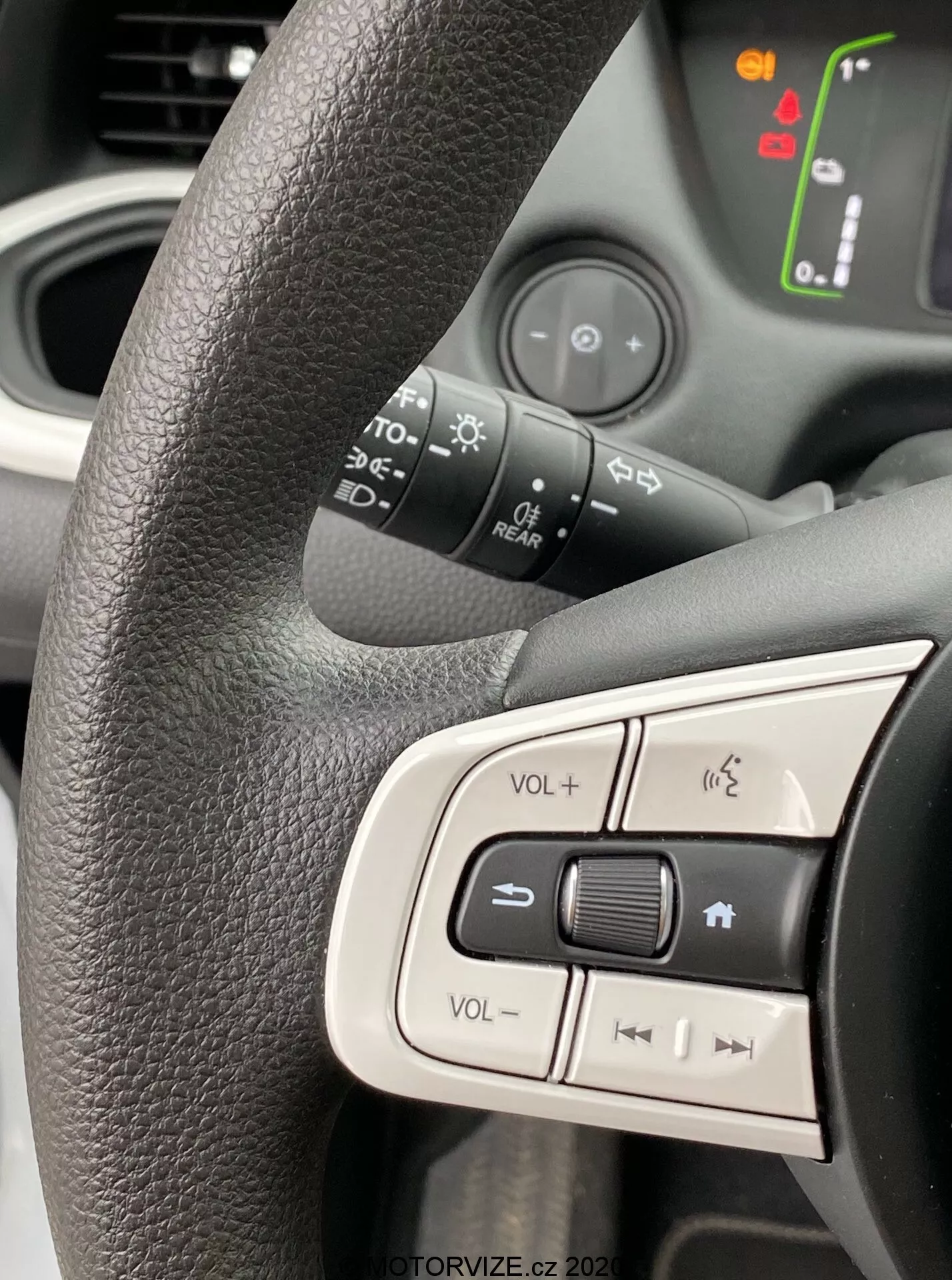 Pohľad zblízka na ľavú stranu volantu Honda Jazz 2020 zameraný na integrované ovládacie tlačidlá. Čierny textúrovaný volant s ergonomickým úchopom, ktorý zobrazuje ovládanie hlasitosti, tlačidlo na stlmenie a tlačidlá na navigáciu v médiách. Mierne rozmazané pozadie odhaľuje osvetlené symboly na prístrojovej doske, čo naznačuje, že vozidlo je zapnuté. Jasné denné svetlo zachytáva moderný, užívateľsky prívetivý dizajn zdôrazňujúci ergonómiu interiéru a technologické vlastnosti.