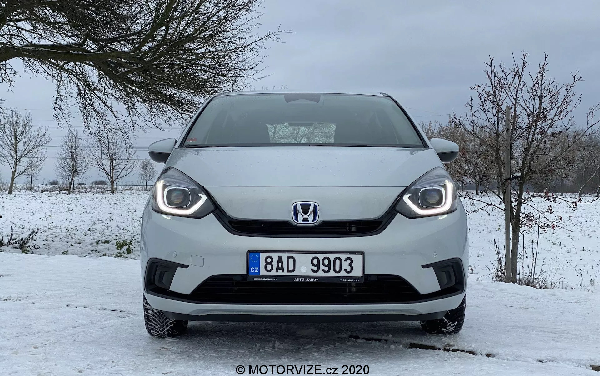 Predný pohľad na Honda Jazz 2020 v zimných podmienkach, ukazujúci výrazný dizajn mriežky, viditeľné svetlomety s LED dennými svetlami a strednú ŠPZ, zaparkovaný na zasneženej ceste s oblačnou oblohou a stromami bez listov.