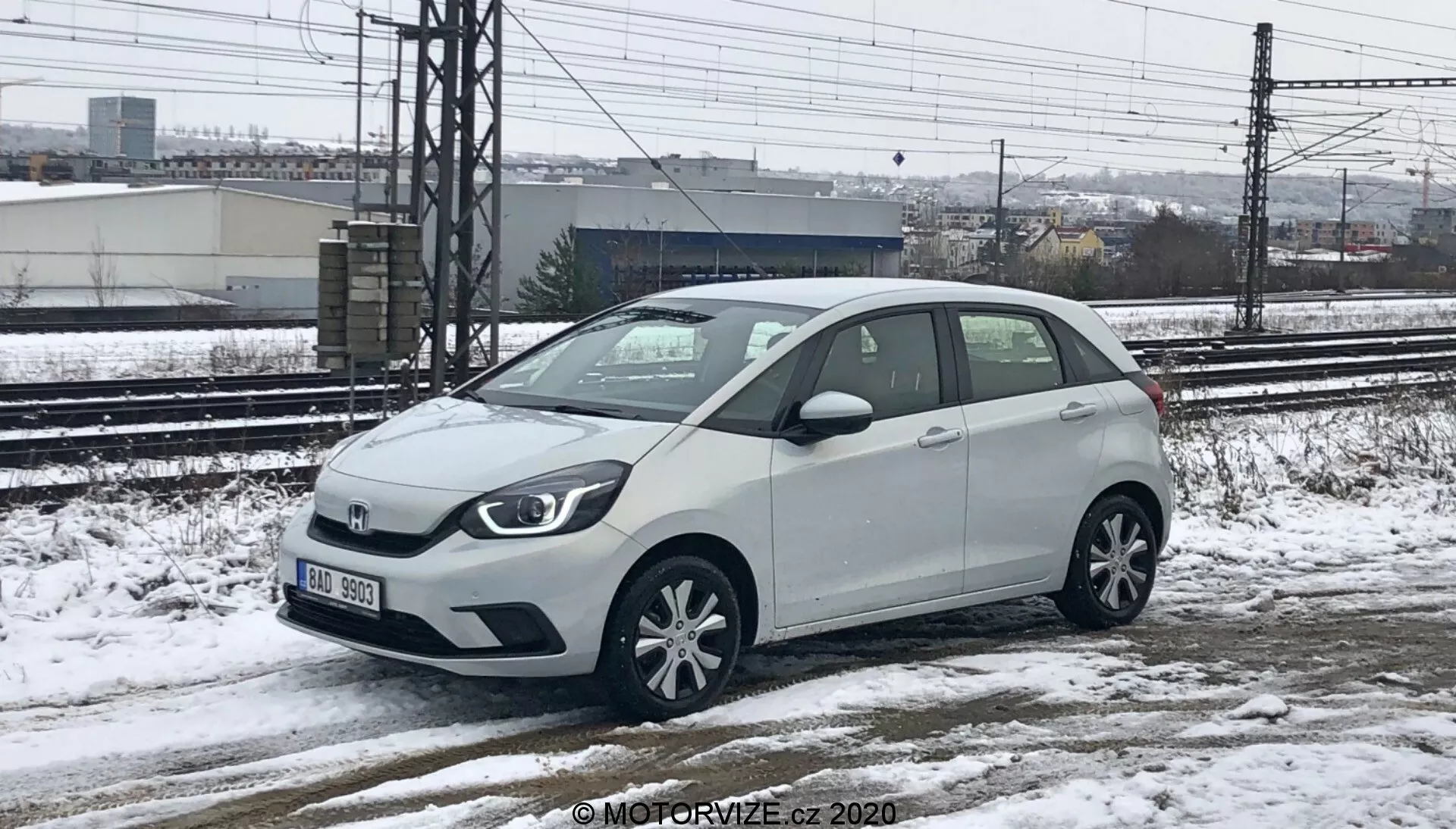 Biely Honda Jazz 2020, subkompaktný hatchback, zachytený z nízkej až strednej výšky, zobrazujúci prednú a pravú stranu. Auto je zaparkované na zasneženom a mokrom povrchu, so snehom viditeľným na vozidle a zemi, čo naznačuje chladné počasie. V pozadí sú viditeľné železničné trate a priemyselné stavby pod zamračenou oblohou. Vozidlo má hlavnú prednú mriežku s logom značky, halogénové svetlomety, viac lúčové zliatinové kolesá, bočné zrkadlá so zabudovanými ukazovateľmi smeru a krátku anténu na zadnej streche.