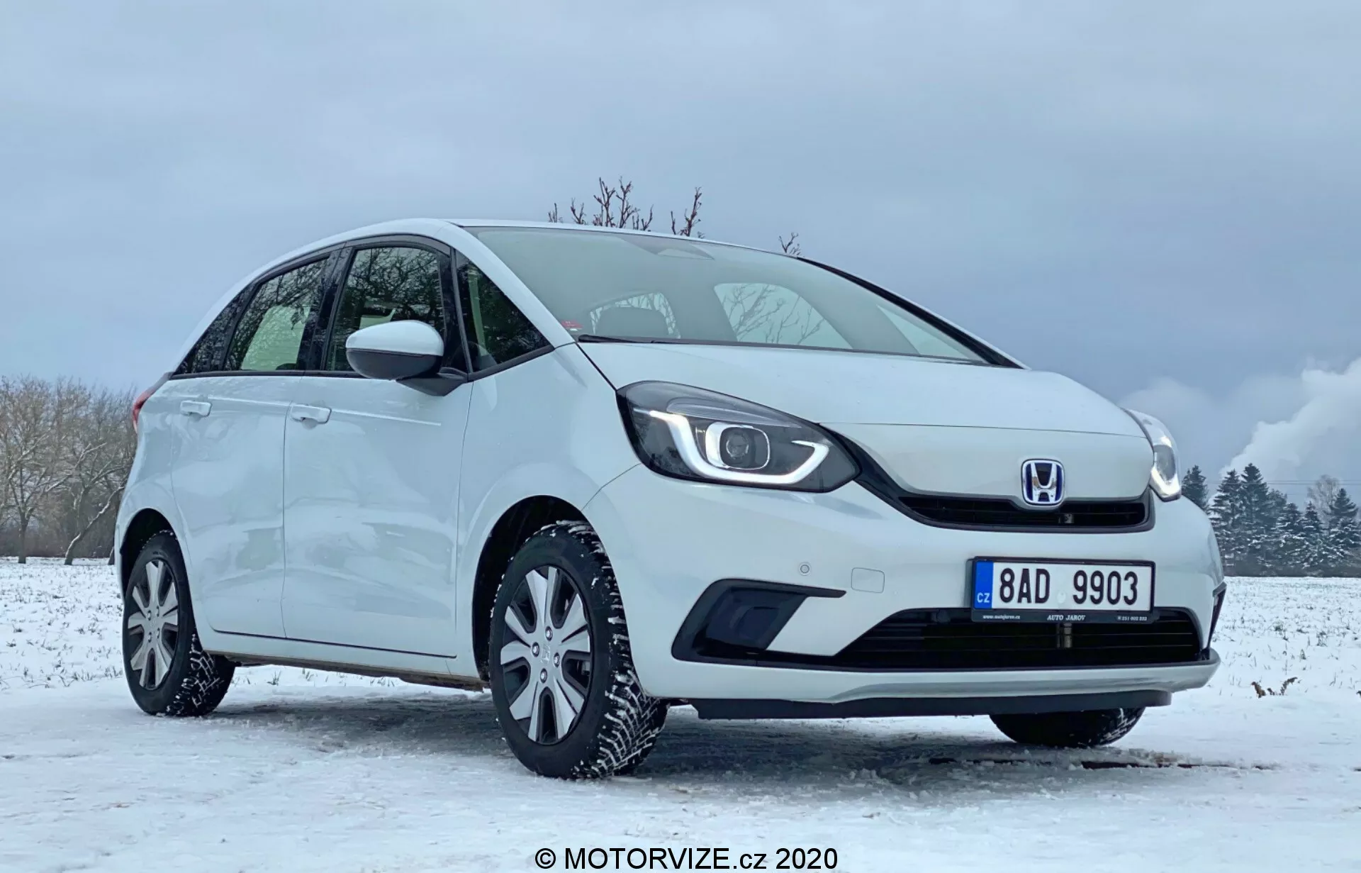 Obrázok zobrazuje biely kompaktný hatchback Honda Jazz 2020 umiestnený v zimnom prostredí so zasneženou zemou a čiastočne zamračenou oblohou. Auto je vidieť z predného štvrtinového uhla zameraného na stranu vodiča, zdôrazňujúc charakteristickú masku chladiča s logom v strede, elegantné LED svetlomety a hladký predný nárazník s integrovanými krytmi hmlových svetiel. Bočný profil ukazuje sklonenú strechu, viacramenné zliatinové kolesá a zrkadlá a kľučky dverí v farbe karosérie. Bezchybný stav vozidla naznačuje, že by mohlo ísť o propagačný alebo recenzný obraz zachytený v roku 2020 MOTORVIZE.cz.