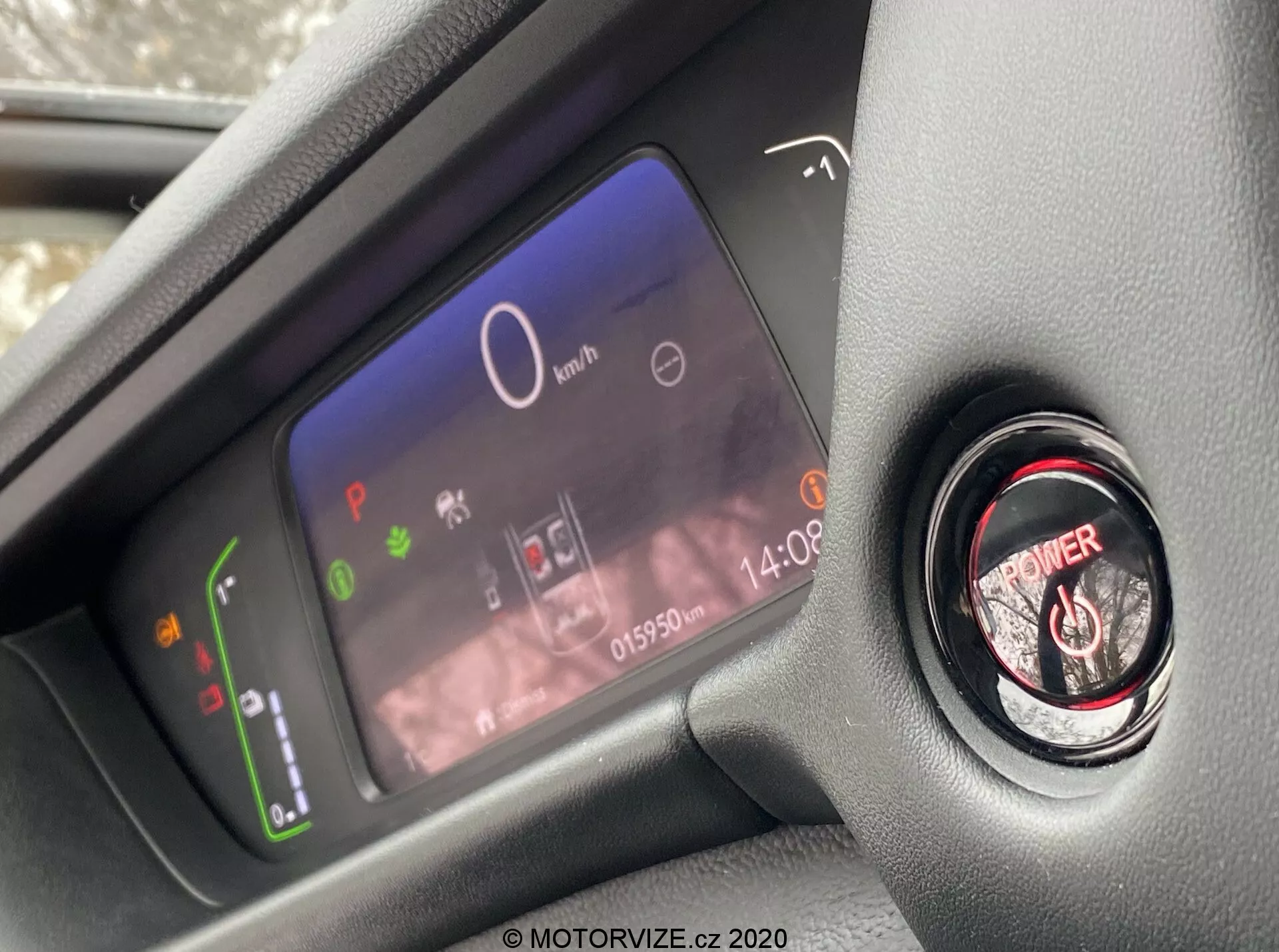 Detailný pohľad na pokročilý digitálny prístrojový panel vodiča a tlačidlo štart/stop motora v modernom interiéry vozidla Honda Jazz 2020, zobrazený z perspektívy sedadla vodiča so zameraním na prvky palubnej dosky. Digitálna obrazovka prístrojového panelu ukazuje výkonový ukazovateľ s ekologickými ikonami, centrálny rýchlomer ukazujúci 0 km/h, rôzne výstražné svetlá, hodiny a odometer s hodnotou 015902 km. Vedľa je tlačidlo zapnutia motora s červeným krúžkom pre bezkľúčové zapínanie.
