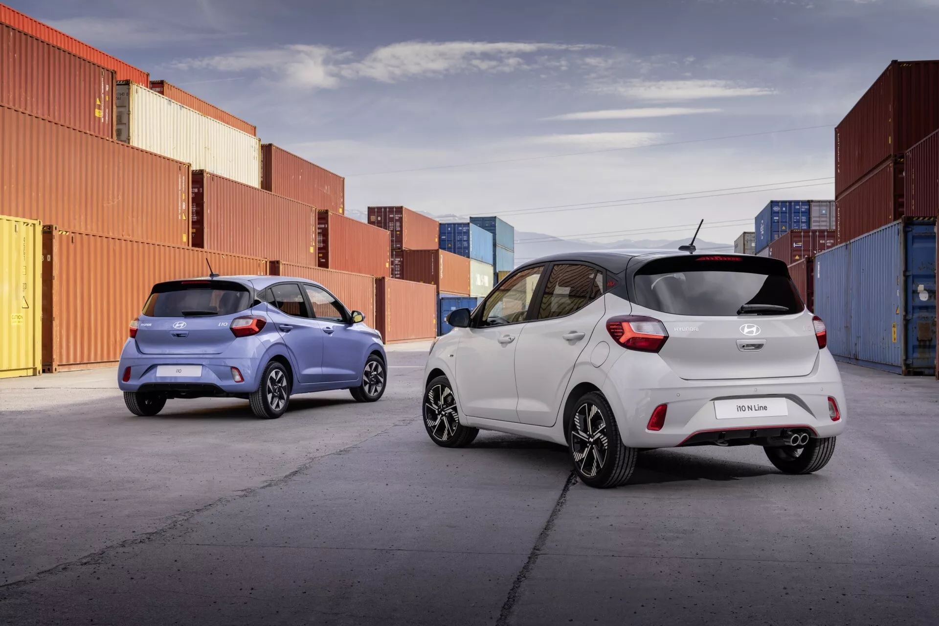 Hyundai i10 2023 (facelift 3. generácie) zobrazené v priemyselnej oblasti, ukazujúce biely a modrý hatchback model s jasnými pohľadmi na dizajnové prvky ako zadný spojler, anténa v tvare žraločej plutvy a kompaktná silueta na pozadí kontajnerov.
