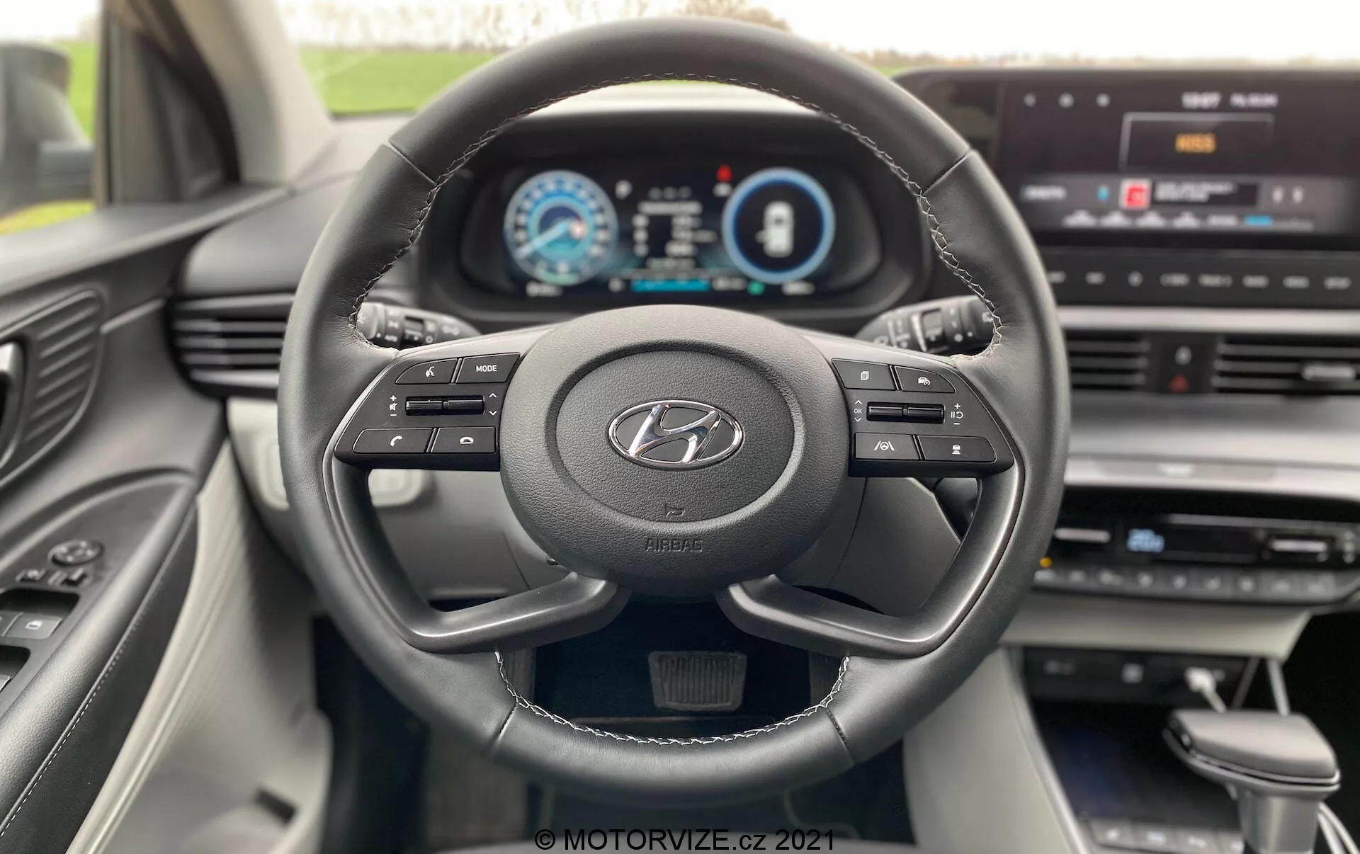 Pohľad na interiér Hyundai i20 z roku 2020 z pohľadu vodiča, zobrazujúci moderný trojramenný volant s logom Hyundai, multifunkčné tlačidlá a plne digitálny prístrojový panel zobrazujúci tachometer, rýchlomer a obrazovku informačného systému vodiča. Palubná doska má centrálny informačno-zábavný displej s možnosťami menu, čo naznačuje pokročilú technologickú integráciu a používateľsky prívetivé rozhranie. Dizajn interiéru je moderný, s čistými líniami a kvalitnými materiálmi viditeľnými v stredovej konzole a dverách spolujazdca.