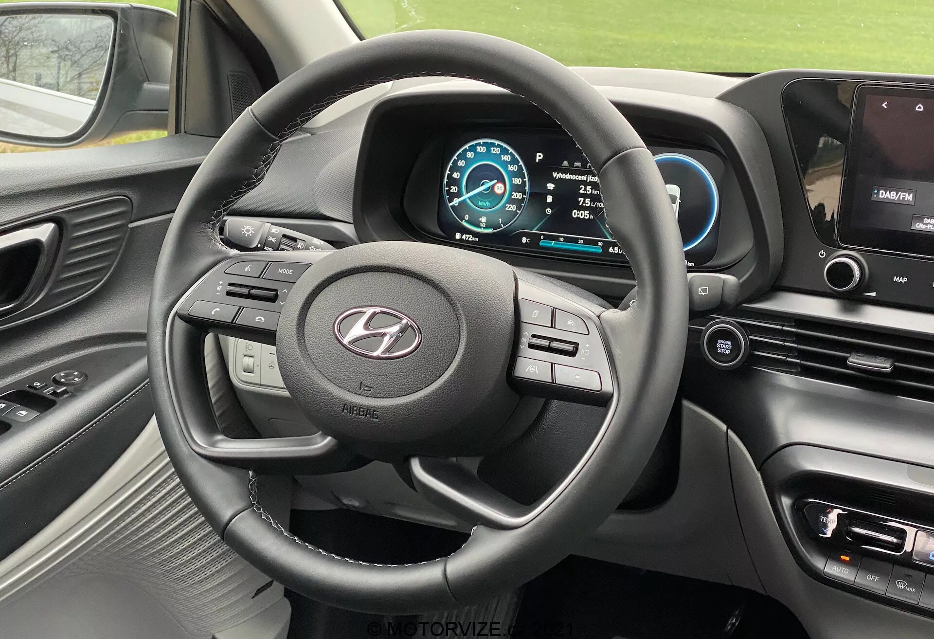 Pohľad na interiér Hyundai i20 2020 z miesta vodiča, zobrazujúci trojramenný volant s viacerými ovládacími tlačidlami, výrazné logo výrobcu, digitálny prístrojový panel s modro-bielou farebnou schémou, tachometer nastavený na 0 km/h, voľbu prevodovky v polohe Park, časť stredového panela s infotainment obrazovkou zobrajujúcou mapu navigácie, ovládacie tlačidlá pre klimatizáciu a nastavenie vozidla, mäkký dotykový dashboard s kovovými akcentami okolo vetracích otvorov a volantu a čiastočný pohľad na vetrací otvor na strane spolujazdca.