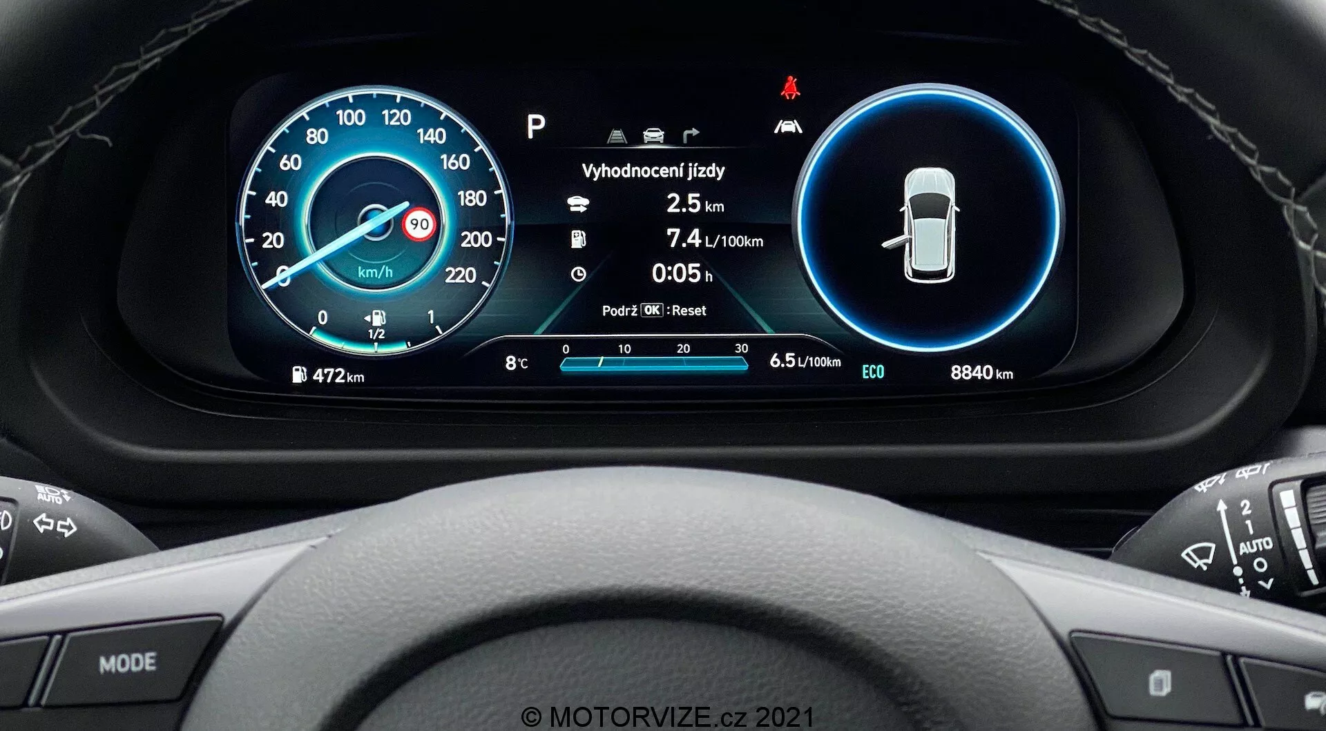 Pohľad z vodičovho sedadla na Hyundai i20 2020, ktorý zobrazuje detailný pohľad na digitálny prístrojový panel umiestnený za volantom. Moderný dizajn obsahuje dva hlavné kruhové displeje: digitálny a analógový tachometer vľavo a ukazovatele bezpečnostných funkcií vpravo. Stred zobrazuje ďalšie informácie, ako sú teplota, počet najazdených kilometrov, spotreba paliva a indikátor výberu prevodového stupňa. Okraj zobrazuje celkový nájazd (8840 km), ukazovateľ paliva a rôzne systémové ikony. Multifunkčný volant s tlačidlami médií a tempomatu je čiastočne viditeľný. V spodnej časti je vodoznak 'MOTORVIZE.cz 2021'.