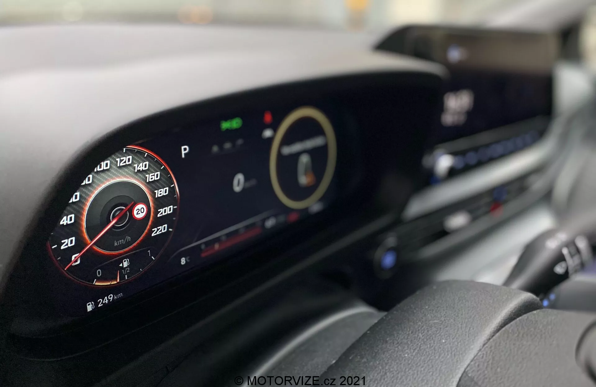 Interiér Hyundai i20 2020 s modernou prístrojovou doskou. Obrázok z miesta vodiča so zameraním na digitálny prístrojový panel a centrálny displej, zobrazujúci detaily ako simulovaný analógový otáčkomer, ekologické indikátory a infotainment systém. Viditeľné sú aj ďalšie funkcie ako vetracie otvory a ovladače klimatizácie.