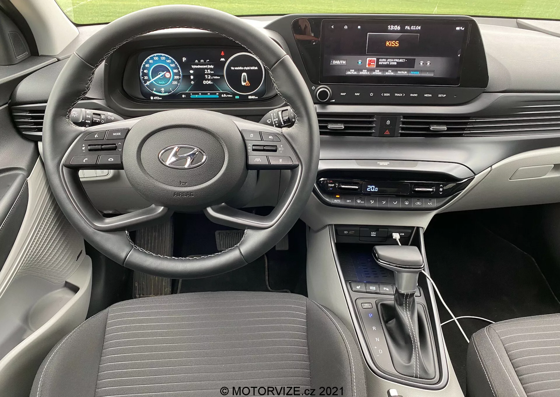 Pohľad na interiér modelu Hyundai i20 2020 z perspektívy vodiča, zobrazujúci trojramenný volant s plochým spodkom a namontovanými ovládacími prvkami pre audio, tempomat a hands-free komunikáciu, plne digitálny prístrojový panel s farebnou grafikou a stredovú konzolu s dotykovým infotainment systémom s multimediálnym pripojením, fyzickými tlačidlami a ovládačmi klimatizácie, modernou radiacou pákou a súčasným vrstveným dizajnom prístrojovej dosky s dvojfarebným schémom.