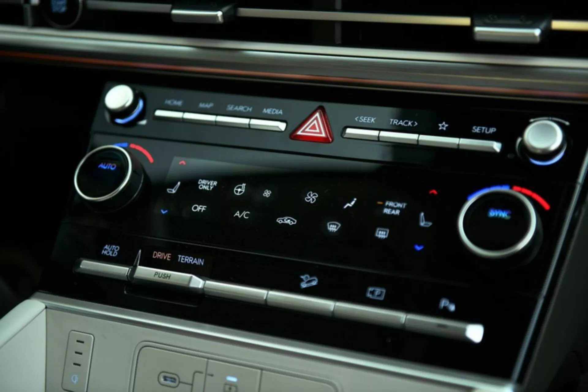 Detailný pohľad zo strany na centrálnu konzolu klimatizácie Hyundai Santa Fe 2024. Obrázok zdôrazňuje dotykové tlačidlá pre ovládanie médií, tlačidlo nouzových svetiel, dvojzónovú klimatizáciu so zobrazenými ikonami, nastaviteľné teplotné kolieska, aktiváciu A/C, ovládanie rýchlosti ventilátora, možnosti jazdných režimov a pohľad do úložného priestoru alebo bezdrôtovej nabíjacej podložky.