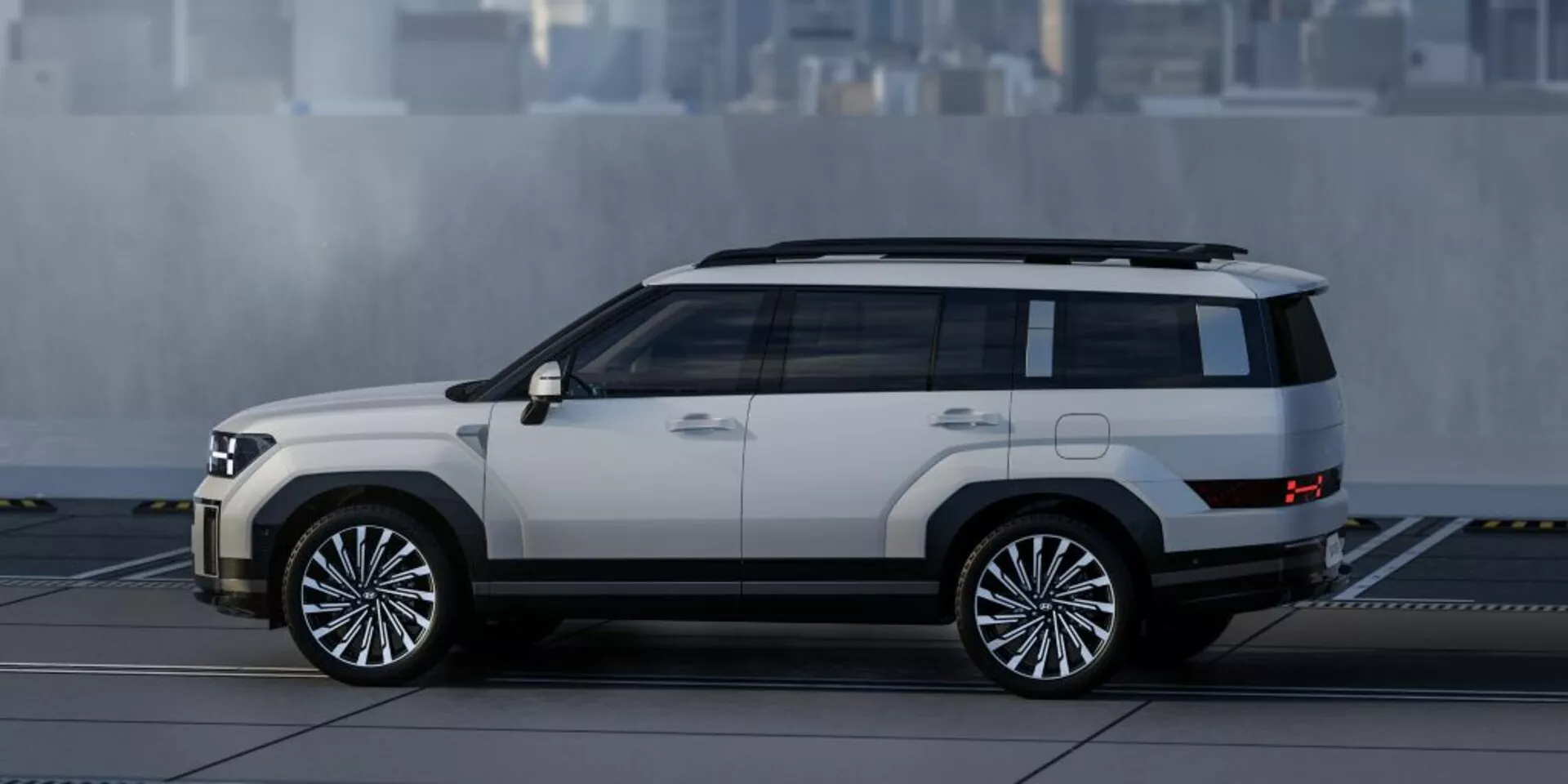 Bočný profil SUV Hyundai Santa Fe 2024, ktorý ukazuje svoj hranatý, ale moderný dizajn s výraznými líniami a hranami. Viditeľné prvky zahŕňajú bočné dvere, okná, bočné zrkadlá, veľké viacramenné hliníkové kolesá, dvojfarebný náter so svetlejšou karosériou a tmavšou strechou a spodným lemom, vysokú svetlú výšku, nášľapy a strešné lišty, všetko na pozadí s rozmazaným mestským prostredím.