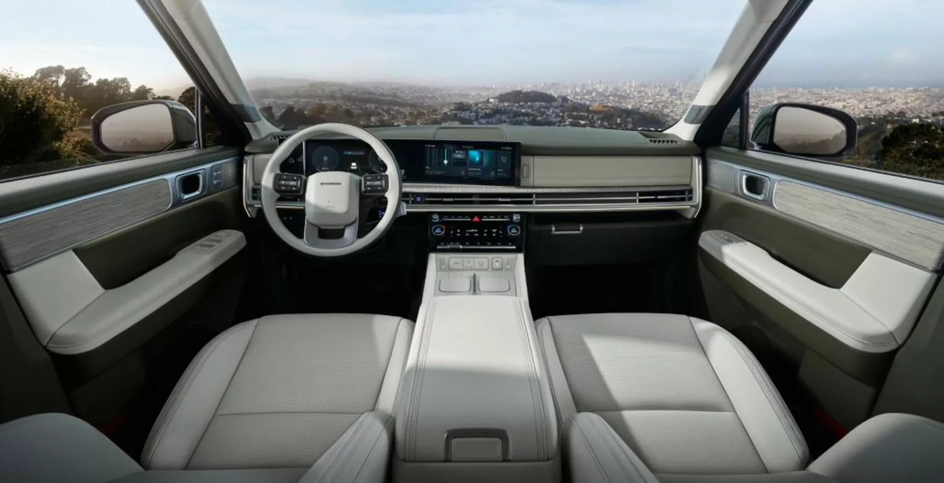 Interiér Hyundai Santa Fe 2024 (5. generácia), ktorý ukazuje prednú kabínu s modernou, minimalistickou prístrojovou doskou, koženým dvojramenným volantom a dotykovou obrazovkou infotainmentu. Stredová konzola obsahuje elegantný elektronický volič prevodových stupňov, držiaky na nápoje a úložné priehradky. Predné sedadlá sú pokryté svetlou kožou, čo zaisťuje pohodlie a ergonómiu. Interiéry dverí zahŕňajú opierky, ovládacie panely pre elektrické okná a ďalší úložný priestor, všetko vyrobené z vysoko kvalitných materiálov. Celkový dizajn vyžaruje luxus, technológie a priestrannosť.