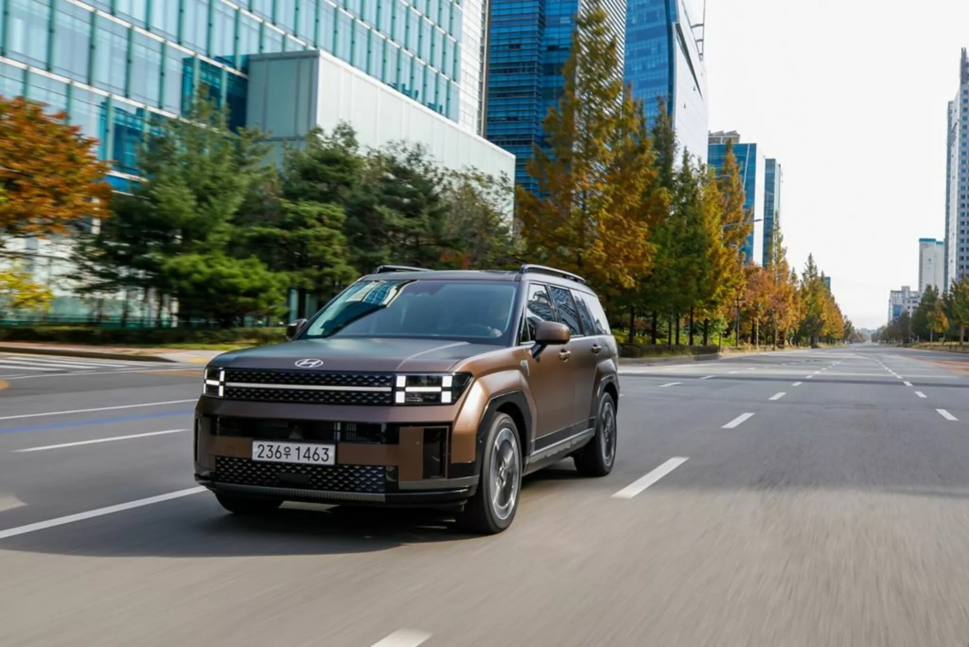 Obrázok modelu Hyundai Santa Fe 2024 piatej generácie, pohľad z troch štvrtín z prednej ľavej strany, zachytený z nízkeho uhlu, ktorý ukazuje elegantnú prednú masku SUV, LED svetlomety, kapotu, predné blatníky, čelné sklo, bočné dvere a bočné zrkadlá. Auto sa pohybuje po mestskej ceste s viacerými pruhmi a komerčnými budovami v pozadí. Aerodynamické kolesá a čiastočne rozmazaná poznávacia značka zvýrazňujú pohyb a výkon vozidla v mestskom prostredí.