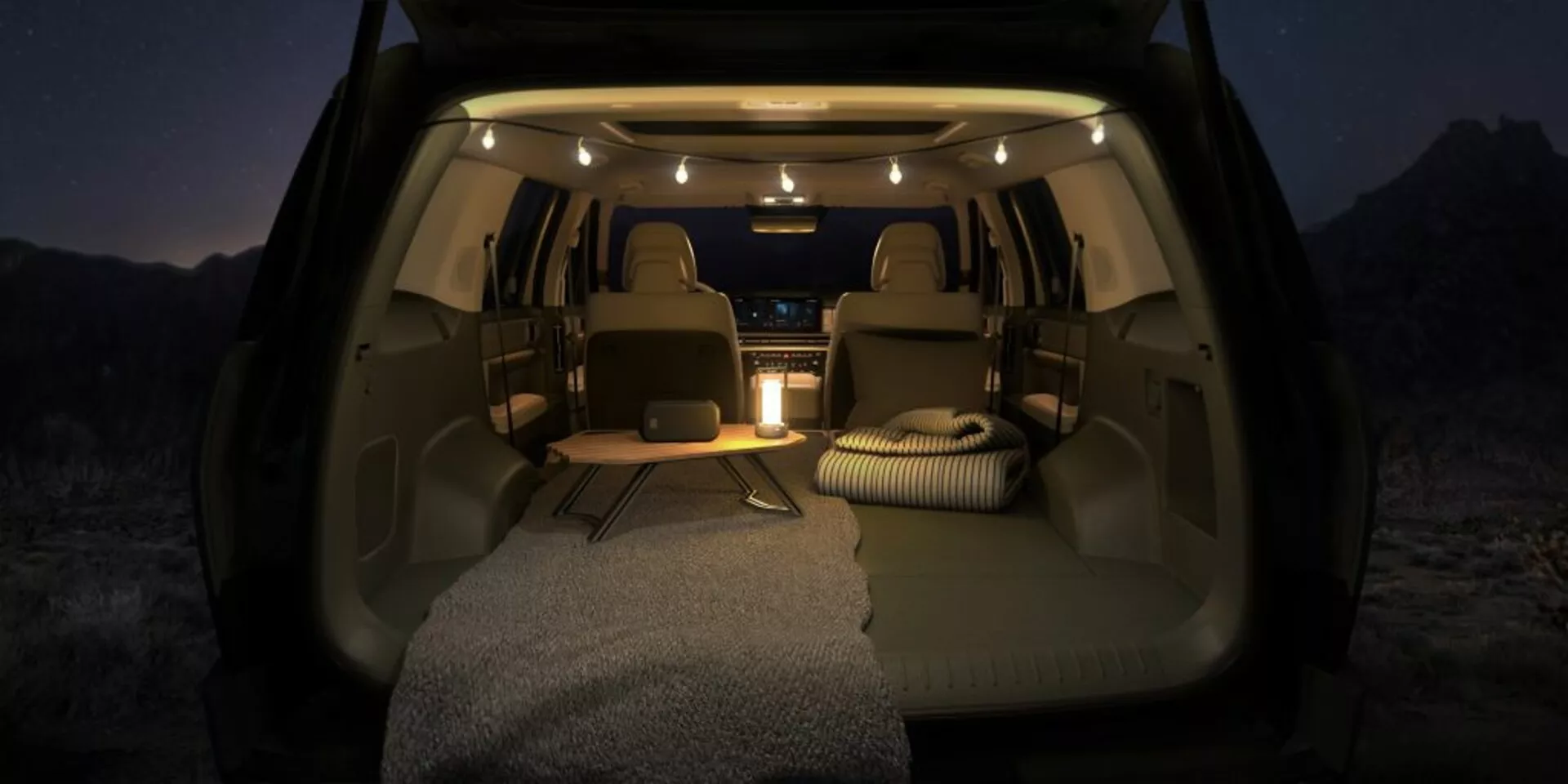 Interiér Hyundai Santa Fe 2024 (5. generácia) z pohľadu zadného nákladového priestoru, ktorý zobrazuje priestrannú a dobre osvetlenú kabínu s sklopnými zadnými sedadlami, vankúšmi a kobercami určenými na rekreačné alebo kempingové použitie. Obrázok zdôrazňuje ambientné osvetlenie, všestranný nákladový priestor s rovnou podlahou a viditeľnú stredovú konzolu s potenciálnymi ovládacími prvkami pre zadných pasažierov a nabíjacími portmi v nočnom prostredí alebo za nízkeho osvetlenia.