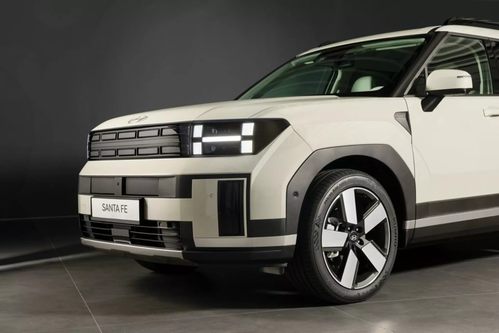 Blízky pohľad z nízkeho uhla na Hyundai Santa Fe 2024 (5. generácia) so zameraním na prednú ľavú štvrtinu, ukazujúci moderný dizajn prednej masky, segmentované svetlomety s charakteristickým denným svietením, sochovanú kapotu, viacúčelové hliníkové koleso, viditeľnú dezén pneumatiky, značkovú masku, zreteľnú líniu na blatníku, aerodynamický predný ľavý dverový panel a zrkadlo a neutrálnu farbu pod štúdiovým osvetlením.
