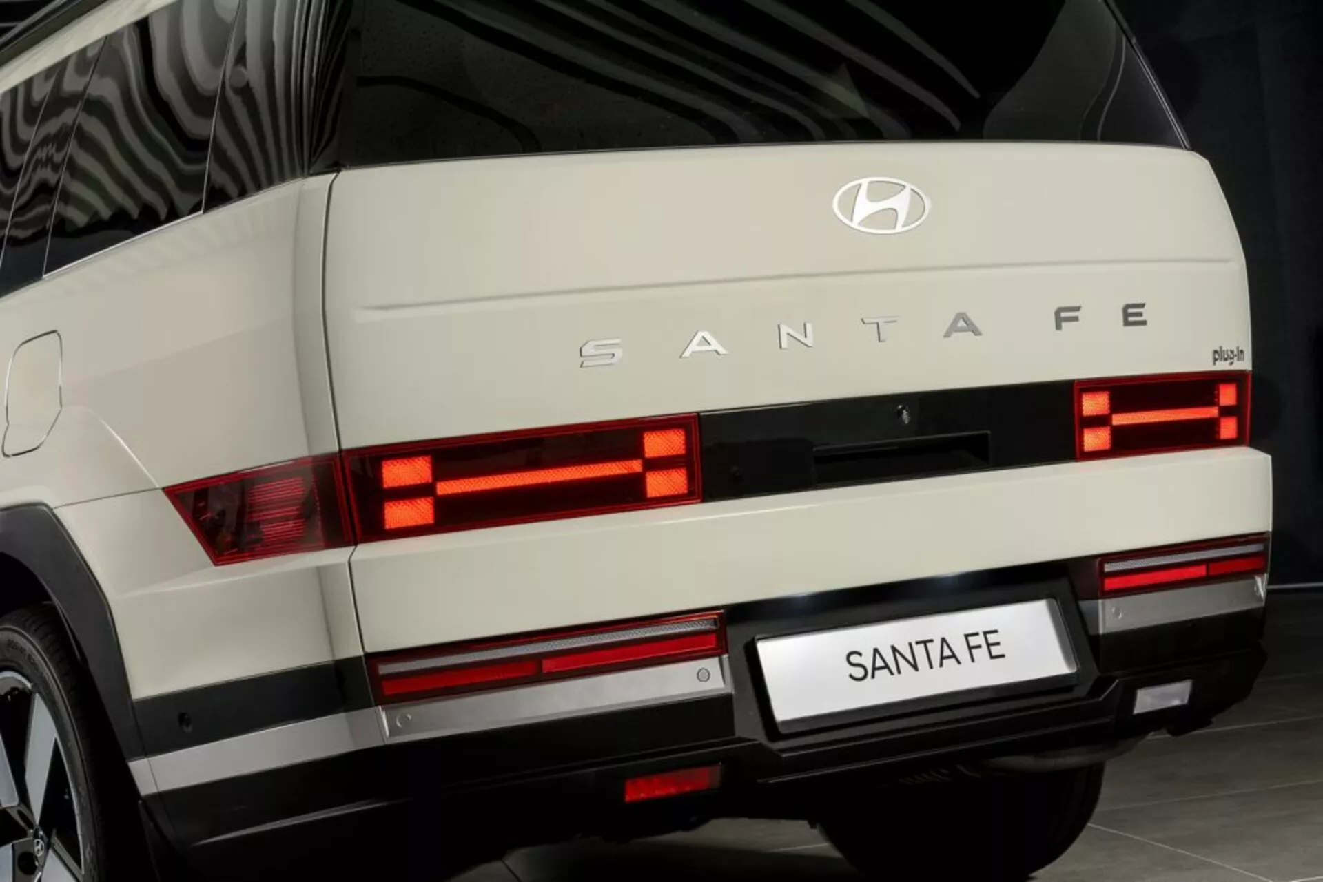 Detailný pohľad na zadnú časť Hyundai Santa Fe 2024 (5. generácia) fotografovaný z úrovne zeme. Obrázok ukazuje osvietené vodorovné segmentované zadné svetlá, strieborný dekoračný prvok, logo Hyundai a označenie 'SANTA FE'. Viditeľné sú zadné stierače, oblasť zadnej poznávacej značky a potenciálne hmlové svetlá alebo odrazky na úrovni nárazníka. Zobrazené v interiérovom prostredí, ktoré naznačuje dobre udržiavané, moderné vozidlo, pravdepodobne použité na propagačné účely.