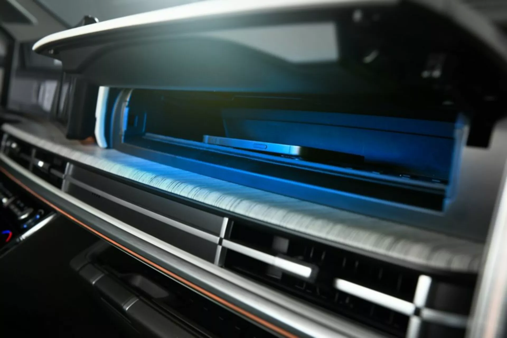 Detailný záber na ventilačný systém vnútri palubnej dosky Hyundai Santa Fe 2024 5. generácie, pohľad mierne zospodu. Horizontálne lamely ventilácie v takmer zatvorenej polohe, s malou hĺbkou ostrosti, rozostrenie popredia a pozadia. Moderný kartáčovaný kovový povrch a potenciálne dotykové ovládanie, zvýraznené modrým ambientným osvetlením pre high-tech, prémiový interiér. Kontrastný materiálový okraj naznačuje luxusnú povrchovú úpravu, možno drevo alebo imitácia dreva. Žiadne viditeľné tlačidlá naznačujú moderný dotykový systém HVAC. Záber interiéru sa zameriava na pohodlie, pohodlie a pokročilé funkcie klimatizačného systému.