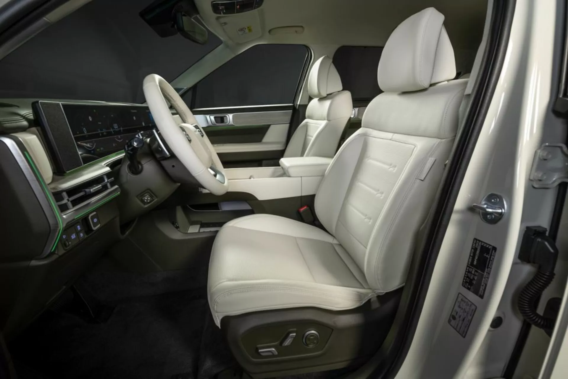 Obrázok zobrazuje luxusný interiér Hyundai Santa Fe 2024 (5. generácia) z vyvýšeného vonkajšieho uhla. Zameriava sa na prednú oblasť vodiča, zahŕňa kožené sedadlo s elektronickým nastavením, panel dverí s ovládaním okien a zrkadiel, multifunkčné volant s funkciami infotainmentu a tempomatu, veľký digitálny displej na prístrojovej doske a imitáciu dreva v interiéri. Stredová konzola naznačuje ďalšie ovládacie prvky a radiacu páku automatickej prevodovky, zatiaľ čo dverový rám zobrazuje informácie o výrobnom procese alebo bezpečnostné údaje.