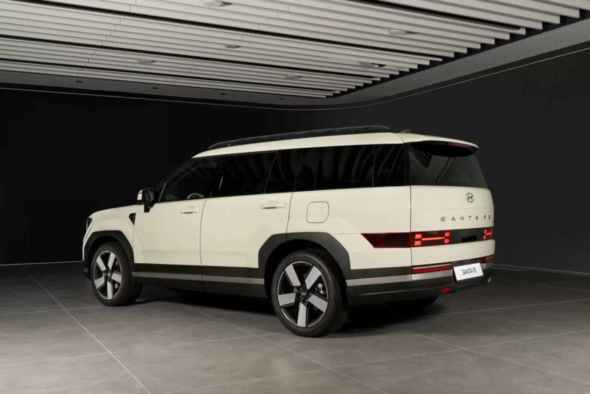 Trojštvrťový zadný pohľad na SUV Hyundai Santa Fe 2024 (5. generácia) zobrazený v neutrálnom vnútornom prostredí. Viditeľné sú zadné svetlá s horizontálnymi čiarami, zadné okno, nárazník a oblasť pre poznávaciu značku. Elegantný bočný profil ukazuje kľučky dverí, obrysové línie a čiastočne viditeľnú panoramatickú strechu. Moderné prvky zahŕňajú veľké zliatinové kolesá s viacerými lúčmi, bočné zrkadlo s integrovanými smerovkami a zatmavené sklá. Svetlá farba SUV a kontrolované prostredie naznačujú, že ide o luxusný model s pokročilou technológiou.