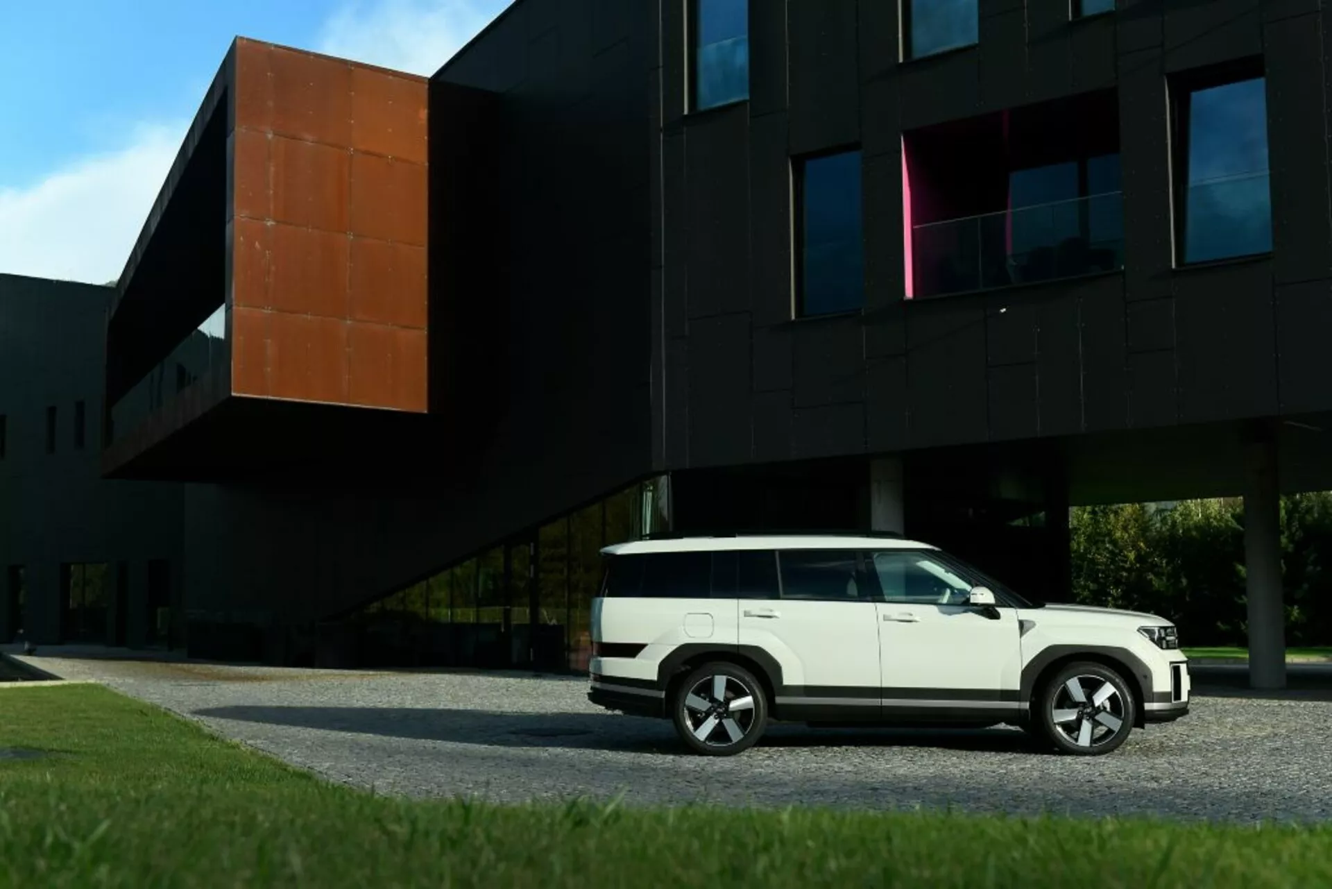 Bočný profil Hyundai Santa Fe 2024 (5. generácia) v bielej farbe, zaparkovaného pred modernou čiernou budovou s oranžovou časťou. Obrázok zobrazuje línie karosérie SUV, bočné dvere, okná, bočné zrkadlá a viacprehohové zliatinové kolesá. Kompozícia zdôrazňuje kontrast medzi moderným dizajnom vozidla a súčasnou architektúrou.