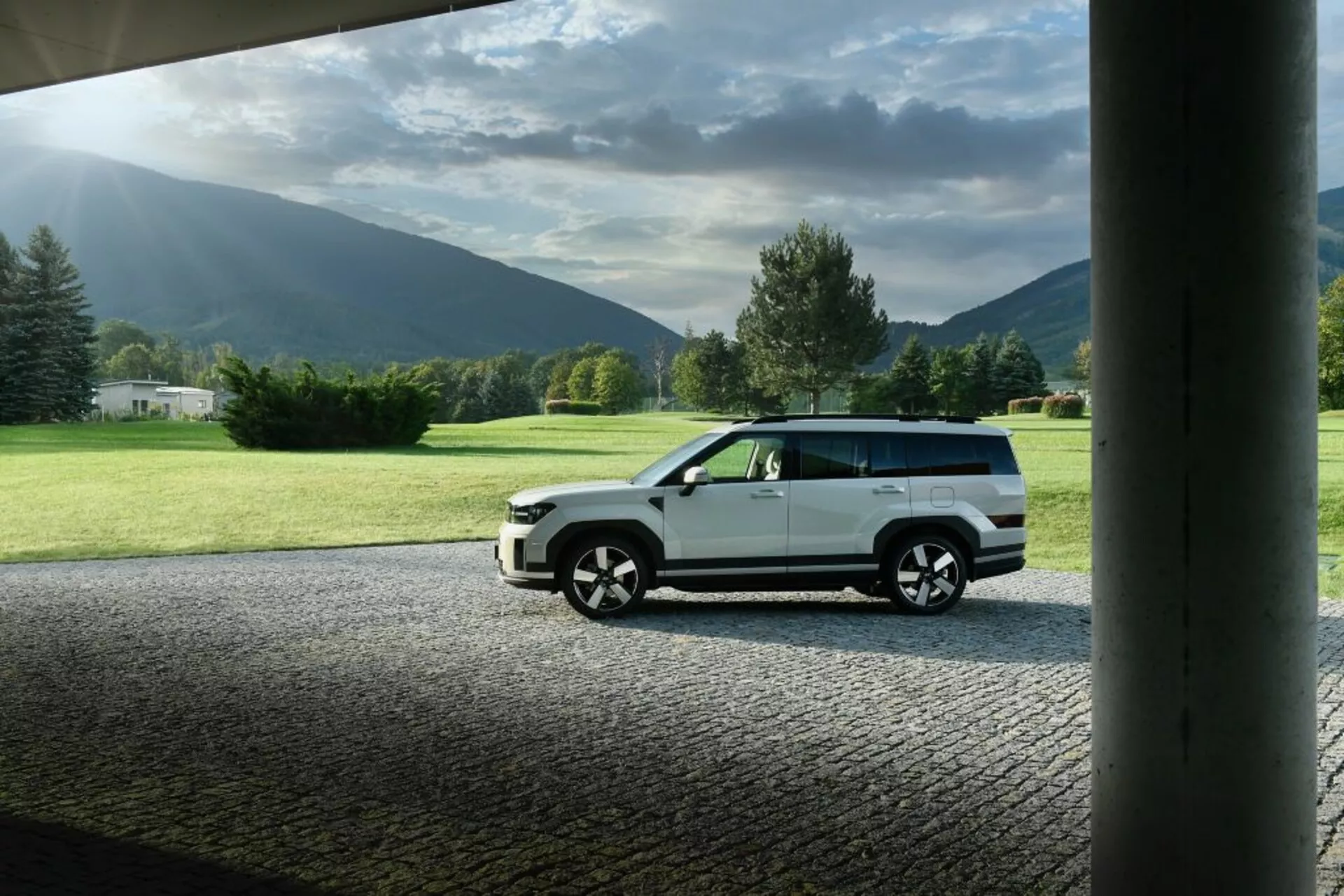 Bočný pohľad na moderné SUV Hyundai Santa-Fe 5. generácie z roku 2024 zaparkované vonku. SUV je umiestnené na spevnenej ploche s trávnatým terénom a horami v pozadí, zachytené z pravej strany na úrovni. Obrázok zvýrazňuje dizajnové prvky vozidla, ako sú línie dverí, bočné okná, veľké zliatinové kolesá a pravdepodobne tmavené okná. Svetlo z ľavej strany vytvára odrazy a tiene, čo naznačuje svetelné podmienky ráno alebo večer.