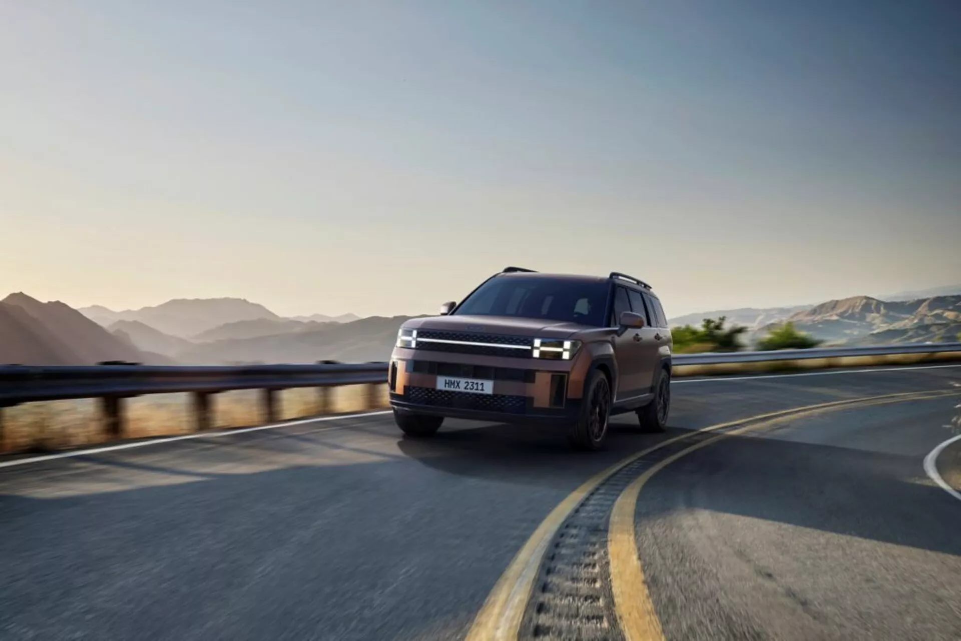 SUV Hyundai Santa Fe 2024 (5. generácia) zachytený v dynamickej perspektíve spredu z troch štvrtín na kľukatej ceste, zvýrazňujúci jeho moderný a elegantný dizajn. Výrazne je zobrazená ľavá strana vozidla, ukazujúca charakteristické LED denné svetlá rámujúce odvážnu prednú masku, úzke svetlomety a viacramenné zliatinové kolesá. Vozidlo je v pohybe, čo demonštruje jeho robustné terénne schopnosti a pokročilé technologické prvky ako ADAS a adaptívne svetlomety, s matným, vysoko kvalitným povrchom.