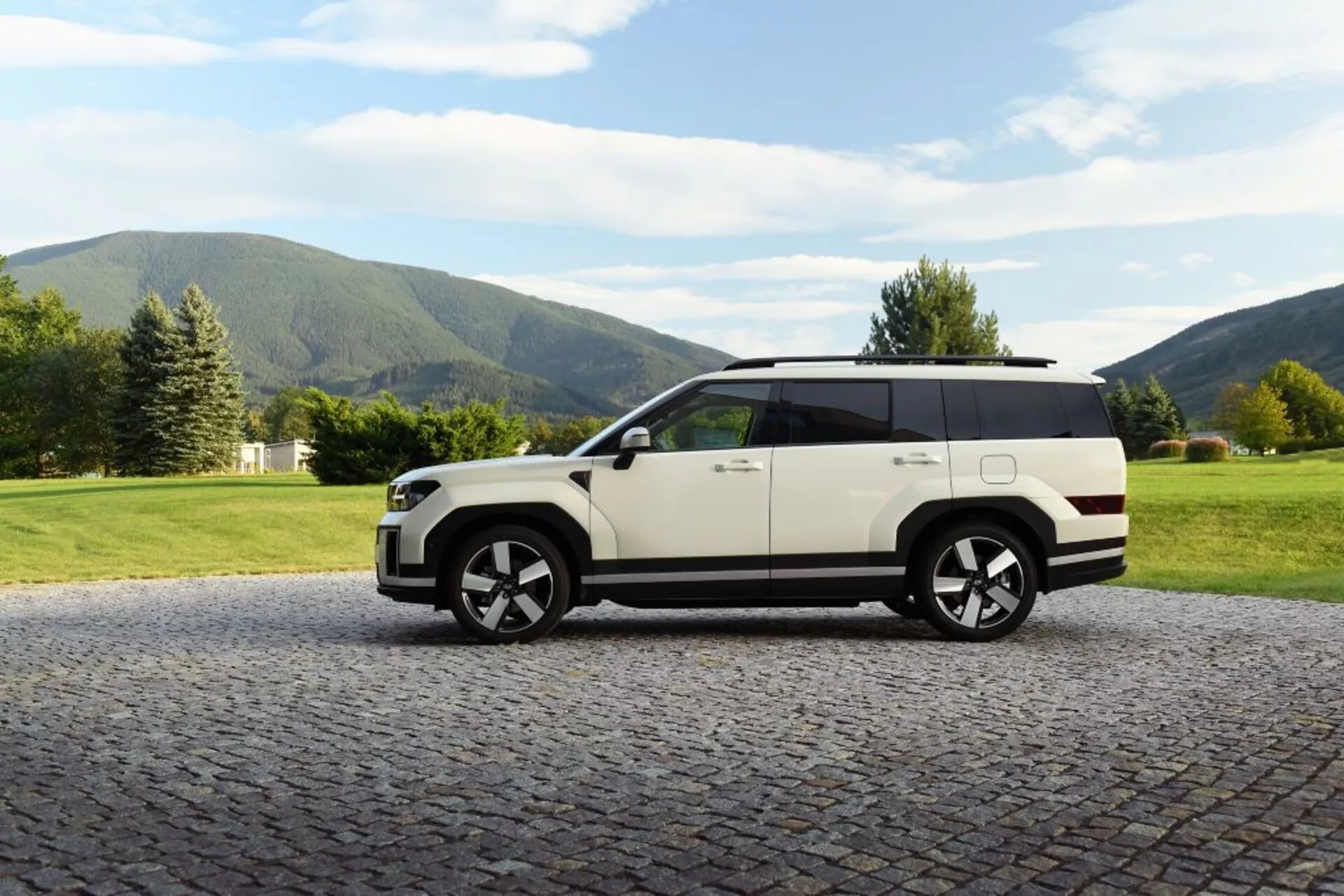 Bočný pohľad na SUV Hyundai Santa Fe z roku 2024 (5. generácia) zaparkované na dláždenom povrchu s prírodným pozadím. Vozidlo má dvojfarebný dizajn s bielou karosériou a čiernou strechou, veľké zliatinové kolesá, viditeľne žiadne kľučky na dverách, čo naznačuje zasúvaciu technológiu, veľké okná, strešné nosiče a vysokú svetlú výšku. Ideálne na predstavenie automobilového dizajnu, luxusu, technologických pokrokov a schopností v teréne.