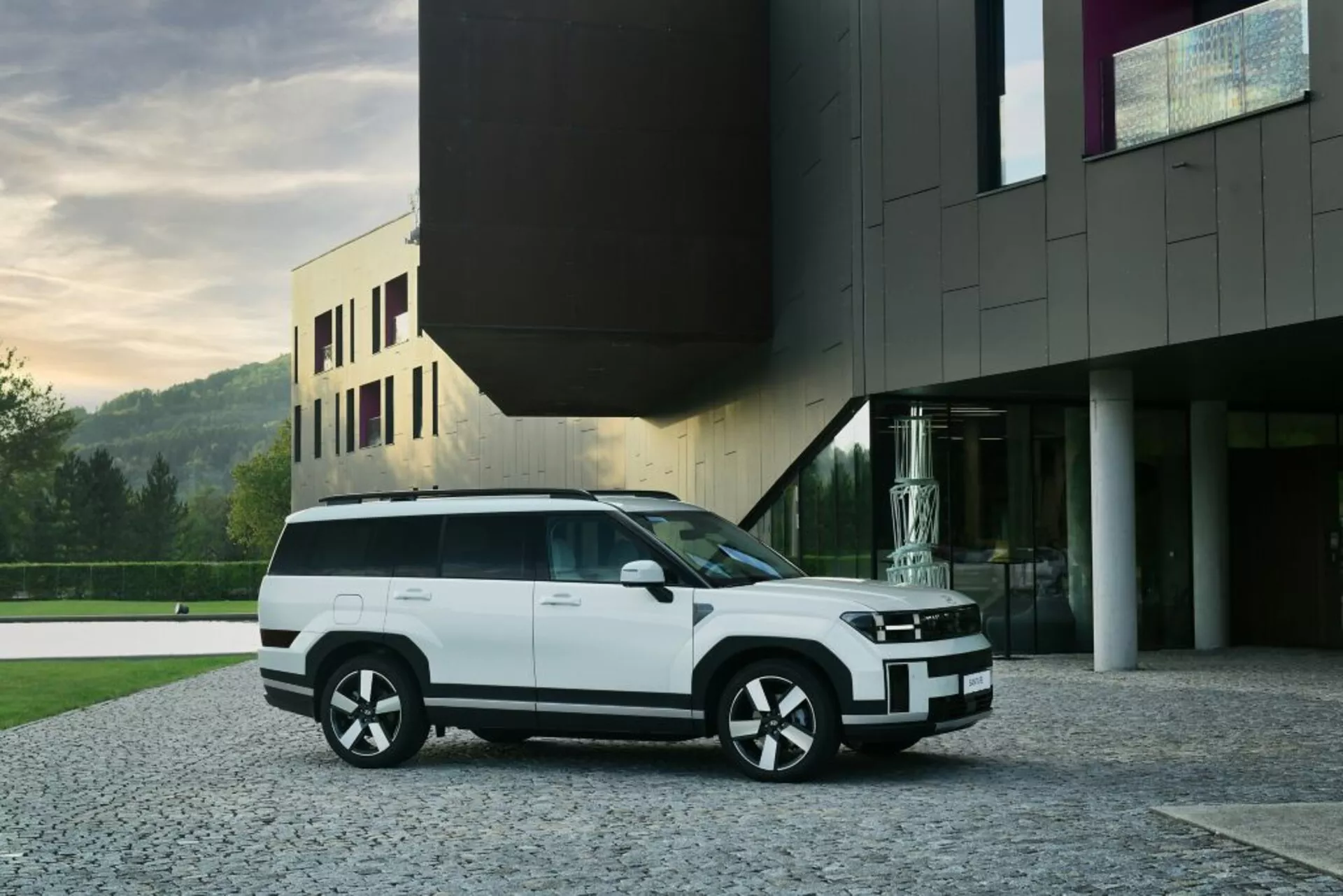Obrázok ukazuje moderné biele Hyundai Santa Fe 2024 SUV (5. generácia), zaparkované na dláždenej príjazdovej ceste pred súčasnou budovou. Zachytené z trojštvrťového predného pohľadu, SUV ukazuje elegantný predný gril integrovaný s horizontálnymi svetlami, nárazník s čiernymi akcentmi a hmlové svetlá. Bočný profil odkrýva veľké okná s čiernymi obrysmi, zapustené kľučky dverí a silnú čiaru ramena. Viacrázové, dvojfarebné kolesá dopĺňajú dizajn vozidla, zatiaľ čo plochá línia strechy s možným panoramatickým strešným oknom a zadným spojlerom zvyšuje aerodynamický štýl vozidla. Obrázok naznačuje LED technológiu v osvetľovacom systéme, čo zvýrazňuje modernú estetiku vozidla. Súčasná architektúra pozadia prispieva k luxusnej prezentácii SUV.