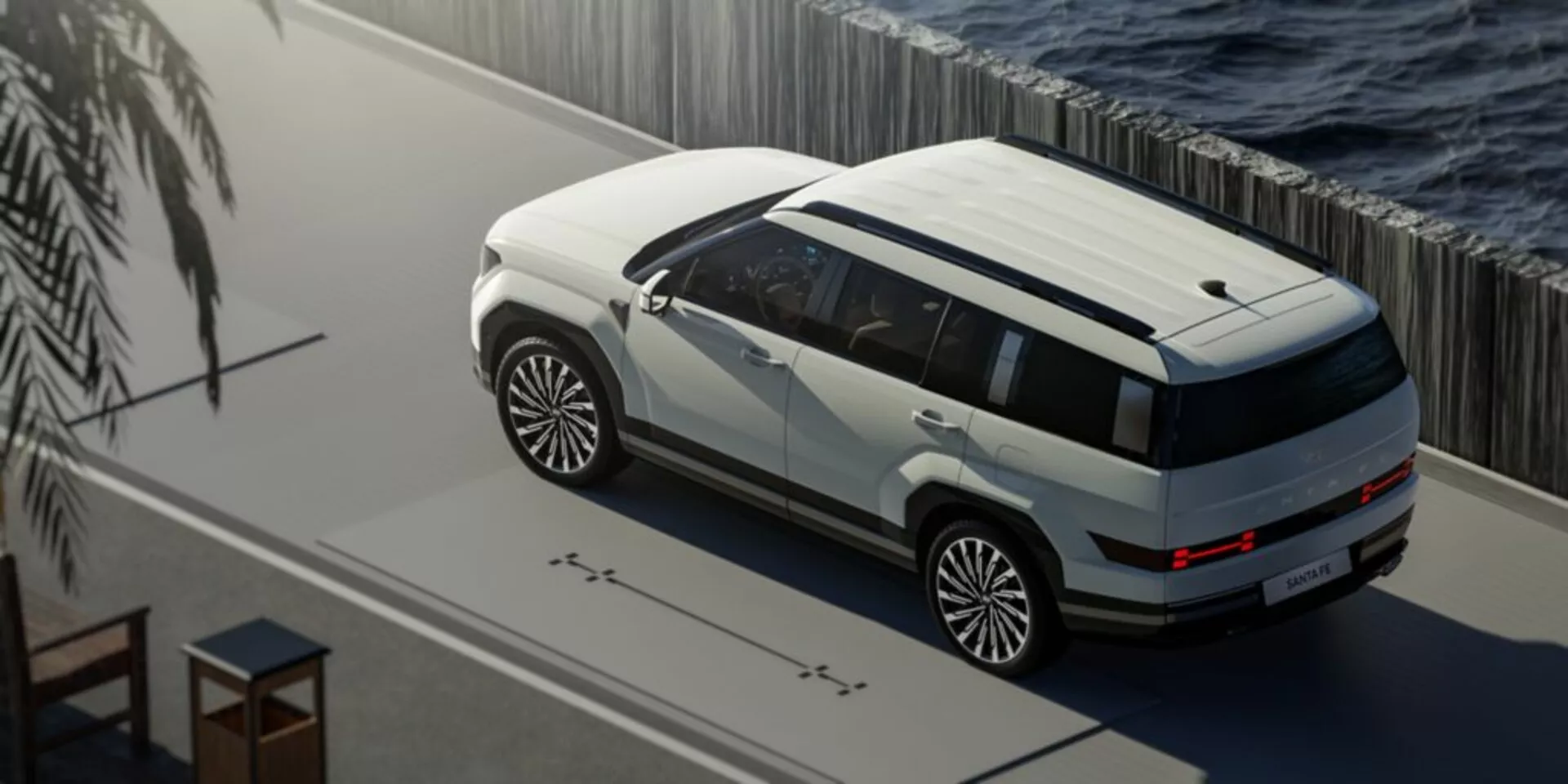 Pohľad zhora na SUV Hyundai Santa Fe piatej generácie 2024 zaparkované pri vodnej ploche, s dvojfarebným lakovaním v bielej a tmavej farbe. Fotografia zobrazuje panoramatické strešné okno, veľké viacramenné disky kolies, karosériovú ochranu a elegantné LED osvetlenie. Scéna je pokojná, s prirodzeným osvetlením naznačujúcim buď ráno alebo popoludnie, a naznačuje luxusné alebo prémiové prostredie s potenciálnymi pokročilými technologickými funkciami, ako sú snímače blízkosti a snímače okolitého svetla.