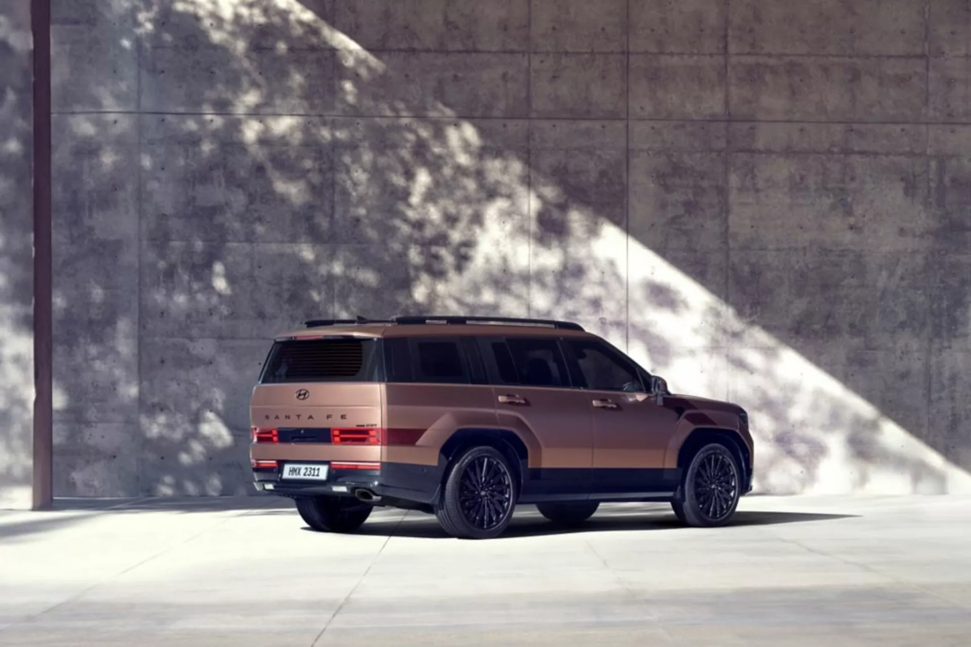Obrázok Hyundai Santa Fe 2024 (5. generácia) v dobre osvetlenom prostredí so silnými tieňmi, zachytený z pohľadu na trištvrte zozadu. Tento moderný SUV je zobrazený z nízkeho uhla, čo zdôrazňuje jeho dominantný a robustný vzhľad. Auto je umiestnené na ľavej strane obrázka, čo naznačuje pohyb smerom doprava. Viditeľné časti zahŕňajú celý profil pravej strany, zadnú časť s výraznými zadnými svetlami, zadný nárazník, spojler strechy a označenie. Vozidlo zaberá značnú časť rámu so svetlom prichádzajúcim zľava, vrhajúc ostré tiene na pravú stranu, čím vytvára prémiový vzhľad s matným bronzovým alebo zemským tónovaným lakom. Mesto alebo architektonické prostredie zvýrazňuje moderný dizajn vozidla. Funkcie, ktoré je možné odvodiť, zahŕňajú zliatinové kolesá, bočné stupienky, strešné okno, LED zadné svetlá a potenciálne pokročilé technológie, ako sú parkovacie senzory a kamery.