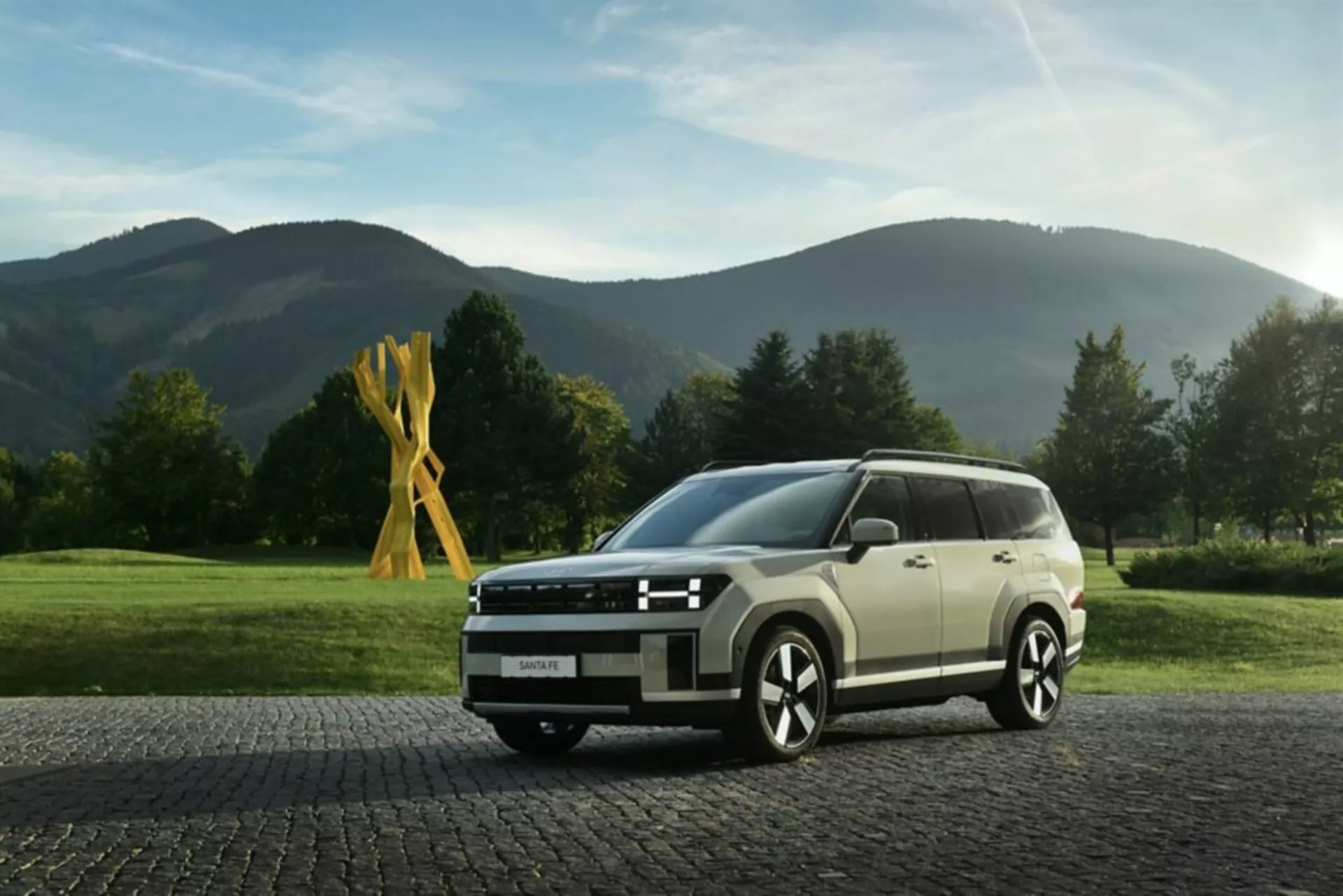 Obraz ukazuje Hyundai Santa Fe 2024, 5. generácie, zachytený v nízkom uhle, z predného kvadrantu, čo zdôrazňuje moderný a hranatý dizajn SUV. Viditeľné prvky zahŕňajú LED svetlomety s ostrým svetelným podpisom, širokú prednú mriežku s horizontálnym pruhovým dizajnom, čiastočne viditeľný nárazník so senzormi alebo kamerami, viaclúčové zliatinové kolesá pre športový vzhľad a bočné zrkadlá s integrovanými smerovkami. Línie karosérie pozdĺž boku zdôrazňujú jazyk dizajnu vozidla, zatiaľ čo veľké okná môžu ponúkať UV filtráciu. Auto je nastavené na dlažobných kockách s pokojným, vidieckym pozadím, čo zvyšuje jeho všestrannú príťažlivosť pre mestské aj vidiecke prostredia. Osvetlenie a hĺbka ostrosti zameriavajú pozornosť na vozidlo, čo ho robí vhodným pre kategórie ako moderné SUV, automobilový dizajn a luxusné vozidlá.