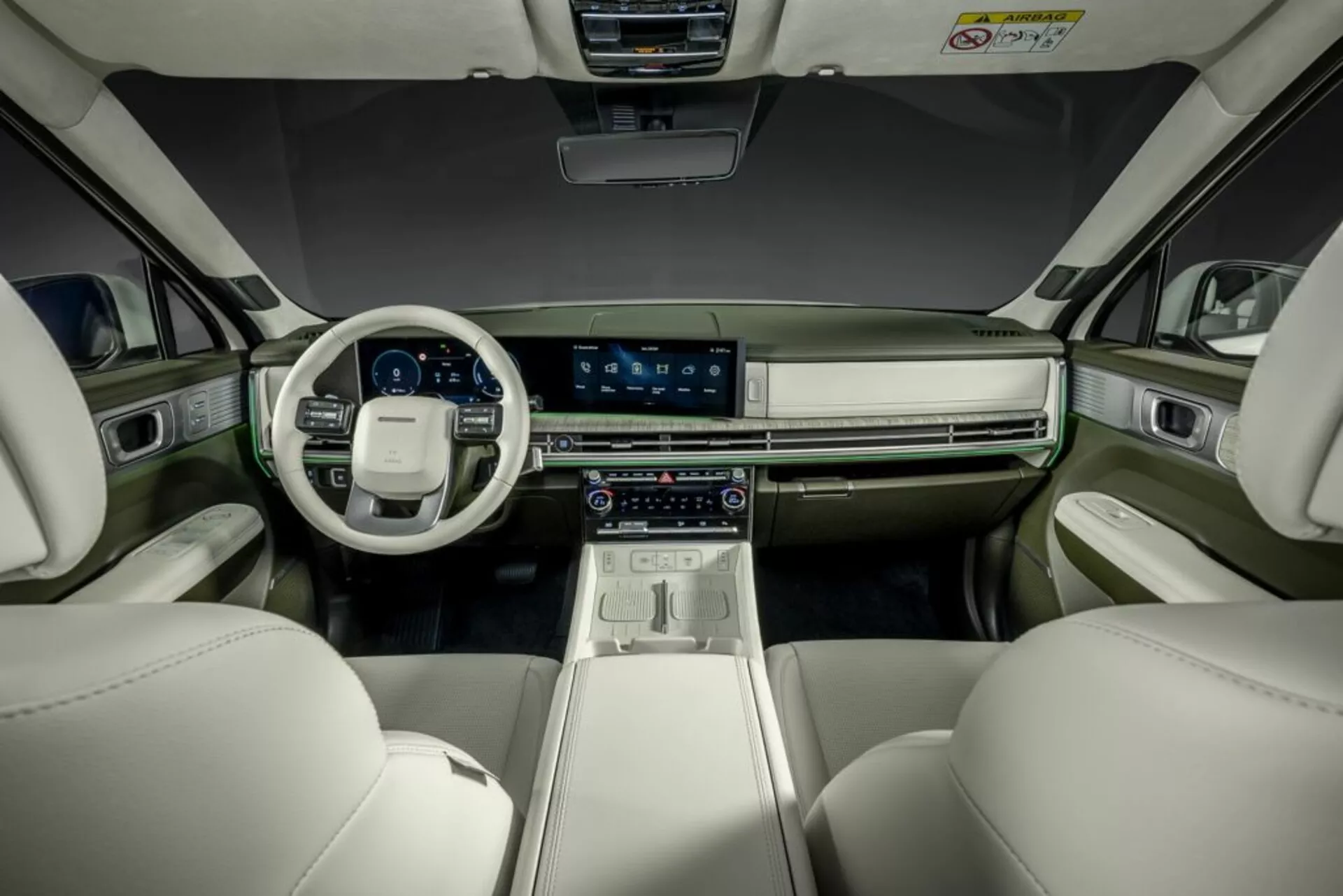 Fotografia interiéru Hyundai Santa Fe 2024 (5. generácia), zachytená zo sedadla vzadu smerom dopredu k palubnej doske. Obrázok zvýrazňuje predné sedadlá a kokpit, s výrazným centrálnym dotykovým displejom, digitálnym prístrojovým panelom, pokročilými ovládaniami klimatizácie a volantom s integrovanými ovládacími prvkami. Moderný, luxusný interiér má dvojtónovú farebnú schému so svetlými koženými sedadlami a tmavšou palubnou doskou a panelmi dverí, zdobenými kovovými prvkami. Ambientné osvetlenie zlepšuje sofistikovanú atmosféru.