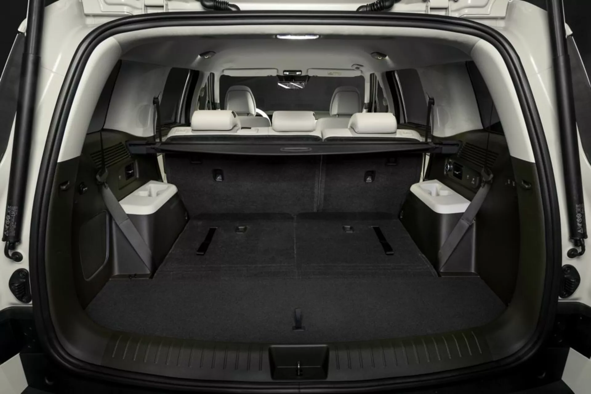 Obrázok zobrazuje interiér nákladového priestoru Hyundai Santa Fe 2024 z pohľadu zozadu, s otvorenými zadnými dverami, ktoré odhaľujú čistú, plochú nákladovú plochu pokrytú kobercom. Tretí rad sedadiel je sklopený na rozšírenie nákladového priestoru, zatiaľ čo stredný rad zostáva vzpriamený pre cestujúcich. Dobre osvetlený interiér zvýrazňuje vlastnosti ako úchytné háčiky, bočné úložné priehradky a integrované panely, ktoré pravdepodobne obsahujú reproduktory alebo vetracie mriežky.