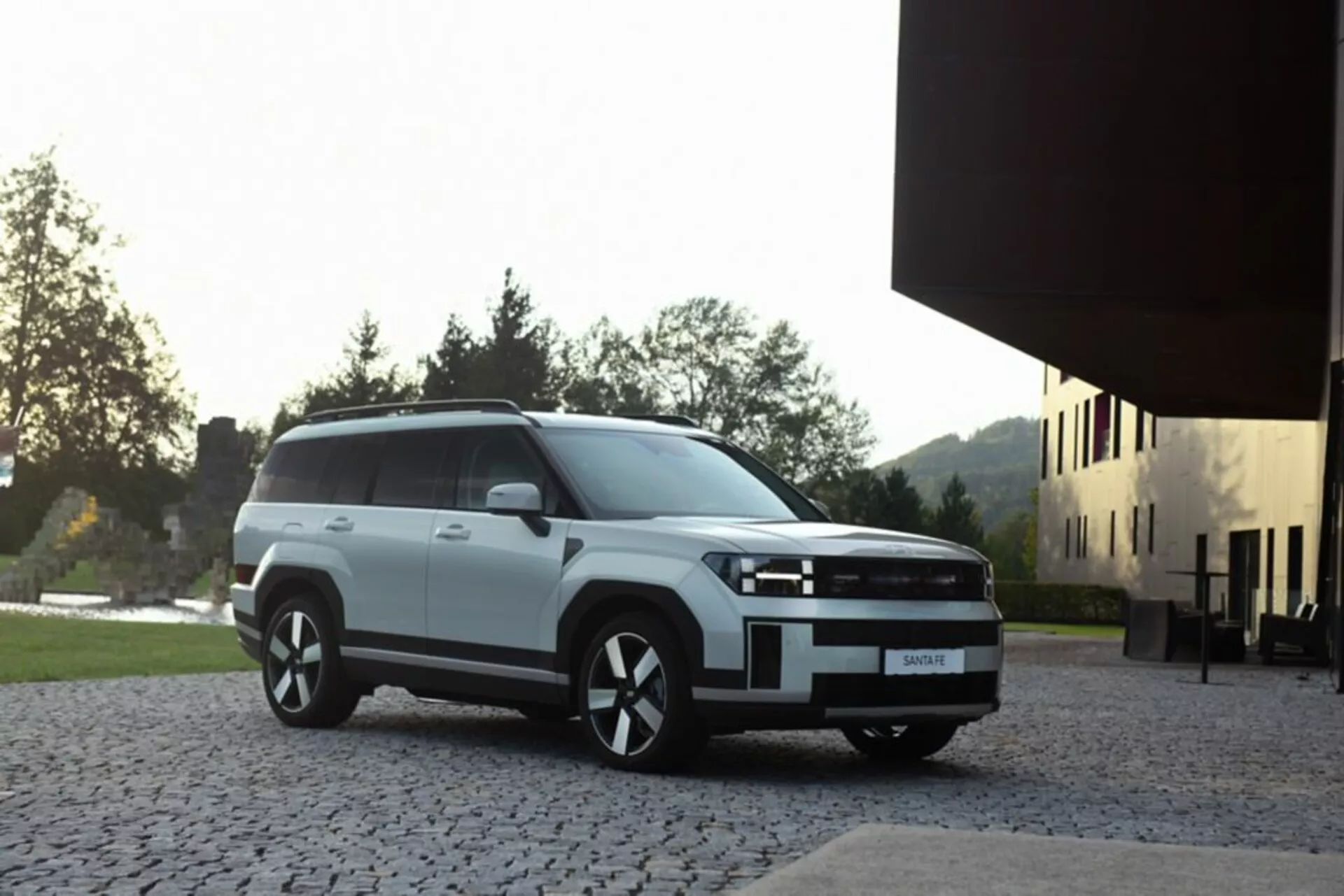 Hyundai Santa Fe 2024 5. generácie v bielej farbe, zaparkovaný na kamennej ploche s modernou budovou a zeleňou v pozadí, zobrazujúci prednú ľavú stranu z trojštvrťového predného uhla, so štvorcovou prednou maskou, LED alebo HID svetlometmi a zliatinovými kolesami, zachytený s jemným prirodzeným osvetlením zvýrazňujúcim modernú estetikou SUV.