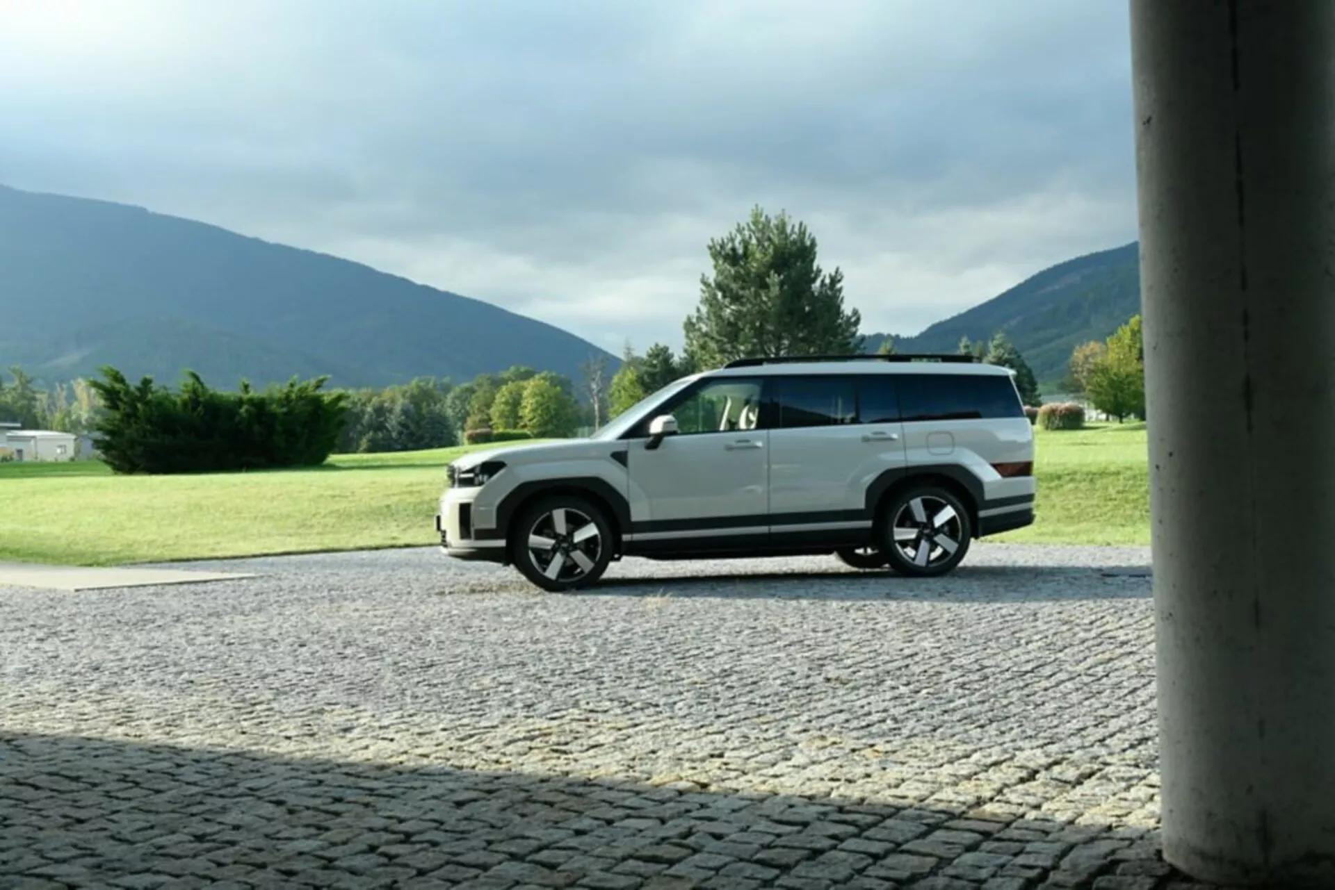 Bočný profil vozidla Hyundai Santa Fe 2024 (5. generácia), zaparkovaného na dláždenom povrchu s prírodným pozadím. Obrázok zachytáva moderný dizajn vozidla, zvýrazňuje veľké viaclúčové kolesá, dvojfarebné lakovanie a detaily karosérie. Fotografia, orámovaná stĺpom budovy, je urobená z nižšieho uhla a zdôrazňuje výšku a postoj auta. Vonkajšie denné nastavenie zvýrazňuje exteriér auta bez silných tieňov.
