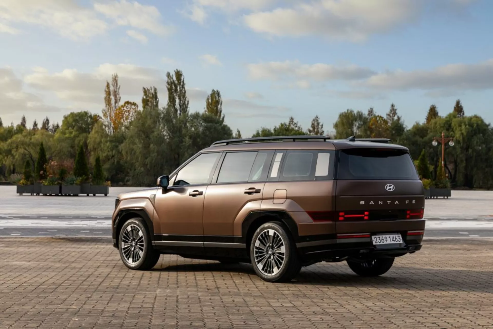 Obrázok zobrazuje moderné SUV Hyundai Santa Fe 2024, 5. generácie, na otvorenom dláždenom priestore obklopenom stromami a modrou oblohou s rozptýlenými oblakmi. Fotografované z uhla tri štvrtiny zozadu, vozidlo zobrazuje vysokú líniu pásu, ploché bočné panely a hranaté blatníky s veľkými, viacramennými zliatinovými kolesami. SUV má dvojfarebný lak v medenej hnedej s čiernou spodnou lištou. Zadná časť obsahuje horizontálne orientované LED zadné svetlá, strieborný ozdobný pásik, označenie modelu 'SANTAFE' a odrazky na nárazníku. Anténa v tvare žraločej plutvy a panoramatická strecha naznačujú pokročilé technologické prvky. Prirodzené denné svetlo zvýrazňujúce kontúry auta dopĺňa scénu.