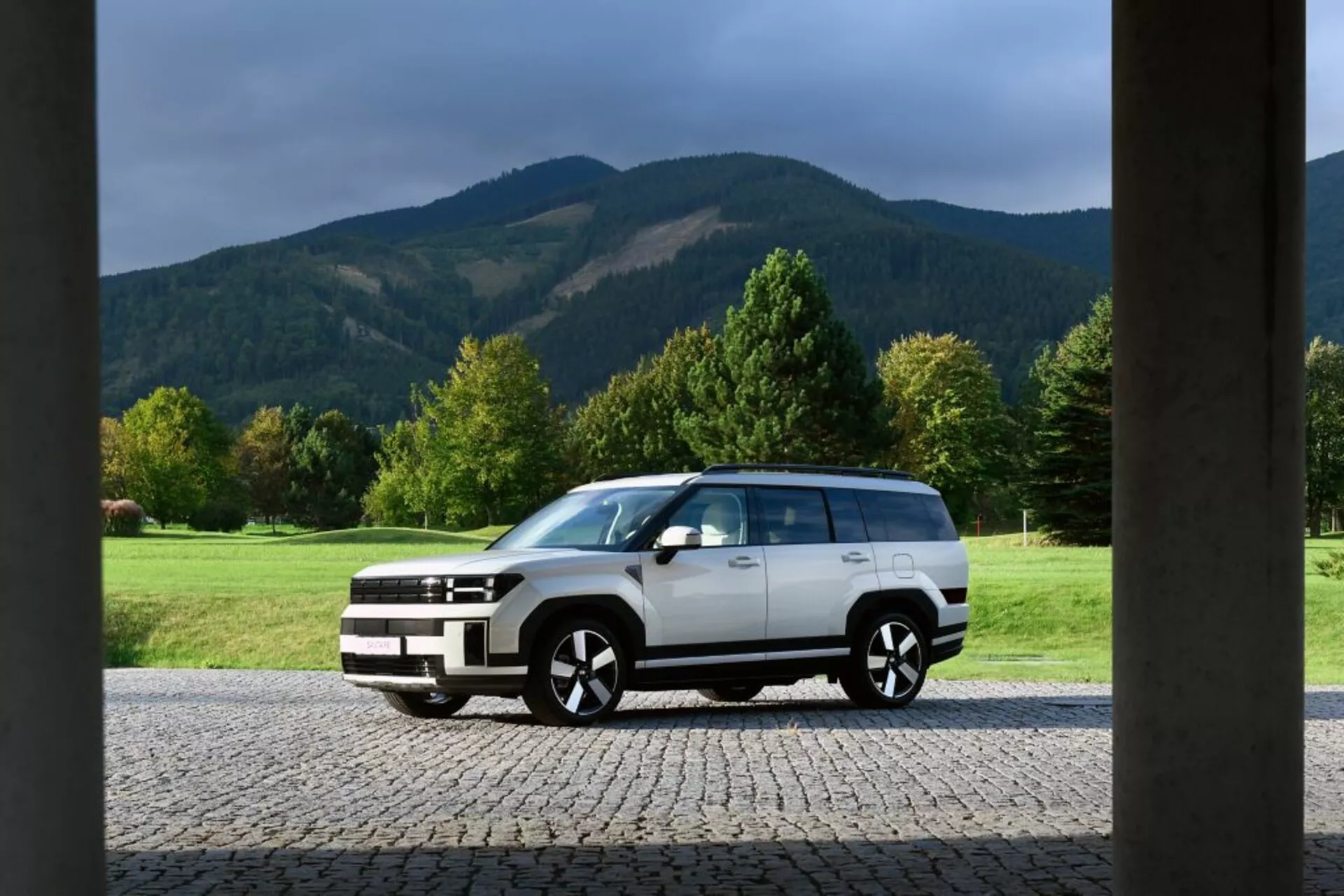 Obrázok predstavuje Hyundai Santa Fe 2024, piatu generáciu, zaparkovanú pred budovou s veľkými stĺpovitými štruktúrami na okrajoch. Auto je plnohodnotné SUV s hranatým, svalnatým dizajnom, ktorý predstavuje modernú estetiku sily a robustnosti. Má dvojfarebné lakovanie s bielym telom a čiernymi lištami na spodnej časti a kolesových oblúkoch. SUV je vybavené veľkými zliatinovými kolesami, tónovanými oknami, LED svetlometmi, dennými svetlami a strešnými ližinami, čo dokazuje jeho pokročilý dizajn a zameranie na úžitkovú hodnotu. Scénické pozadie zahŕňa otvorené trávnaté pole, stromy a hory, zdôrazňujúc dobrodružné vonkajšie prostredie.