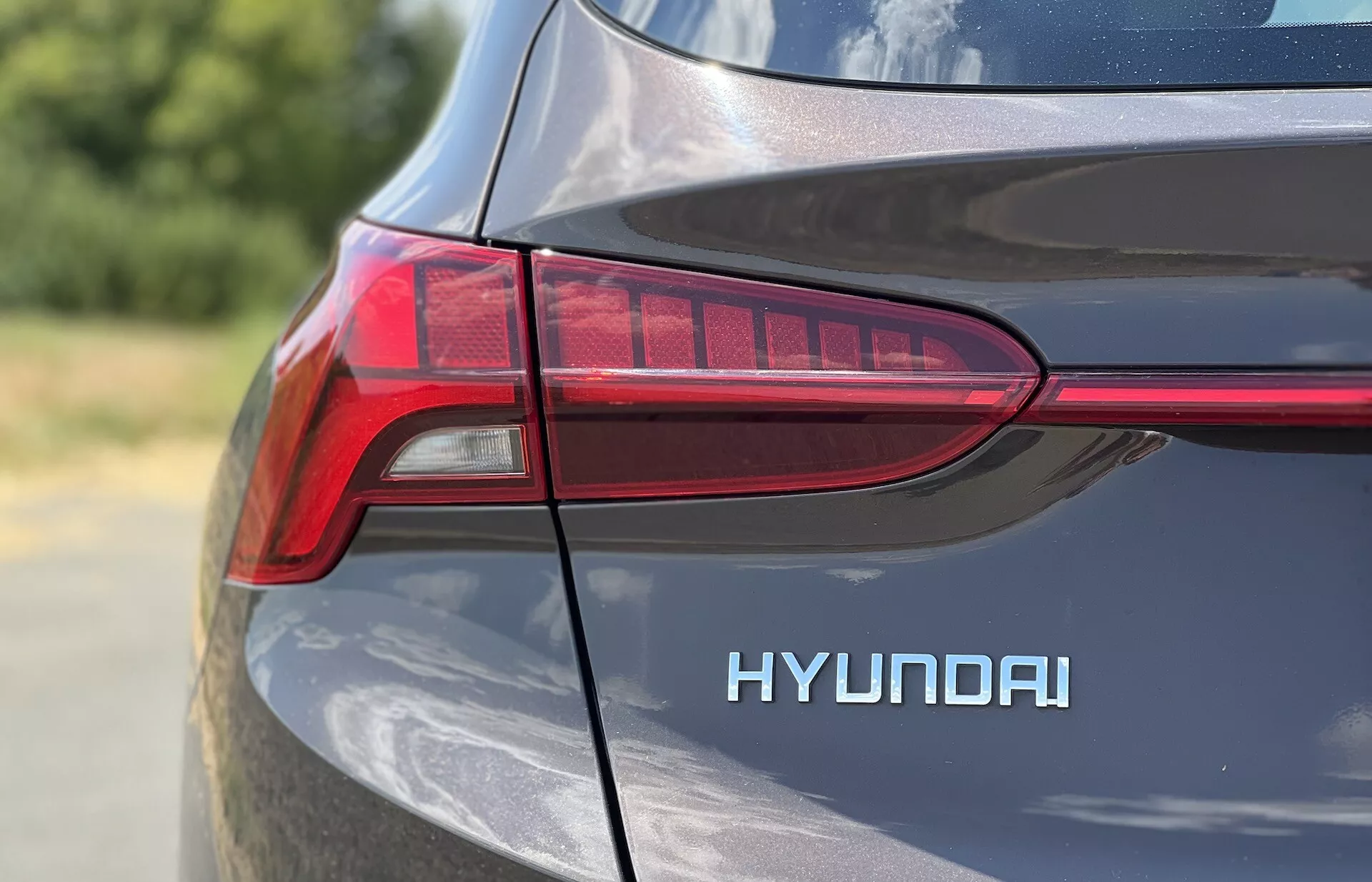 Detailný záber na zadnú štvrtinu na strane vodiča Hyundai Santa-Fe 2020, so zameraním na LED svetlo v tvare T a výrazný nápis 'HYUNDAI'. Kovový lak odráža denné svetlo a zdôrazňuje krivky a línie dizajnu v tesnom, detailnom zábere s malou hĺbkou ostrosti.