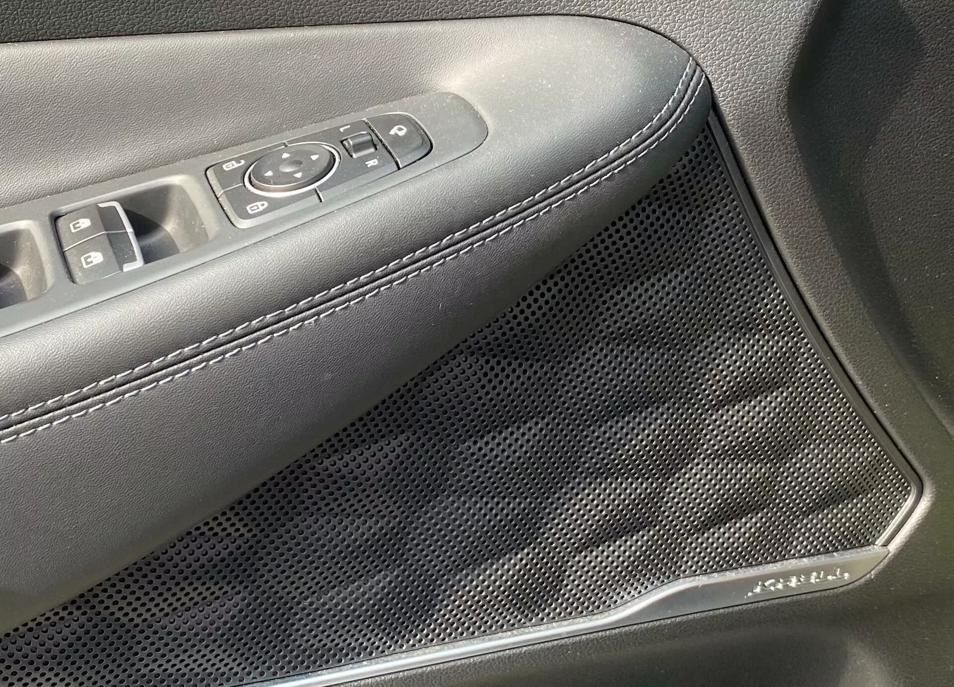 Detailný pohľad na vnútorný panel dverí Hyundai Santa Fe 2020, ktorý zvýrazňuje oblasť s ovládaním elektrických okien a zrkadiel a tiež mriežku reproduktora so vzorom včelieho plástu. Opierka ruky je čalúnená tmavým materiálom s bielymi švami. Mriežku reproduktora zdobí dekor pripomínajúci chróm, čo naznačuje pokročilé audio a estetické dizajnové prvky.