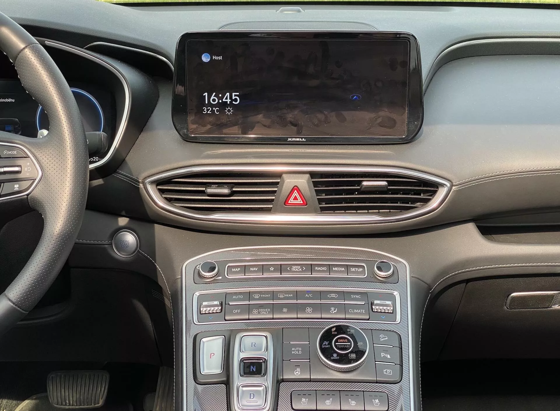 Blízky, čelný pohľad na centrálnu konzolu a časť palubnej dosky moderného auta, vrátane infotainment displeja ukazujúceho 16:45 a 32°C, aerodynamické ventilačné otvory, tlačidlo výstražného svetla, kombinácia dotykových a fyzických tlačidiel s kovovými akcentmi, otočný ovládač na riadenie infotainmentu a navigácie a volič jazdných režimov, čo naznačuje elegantný a moderný interiér s pokročilými funkciami, ako je klimatizácia, vyhrievanie/chladenie sedadiel a rôzne jazdné režimy. Rozmazaná palubná doska a čiastočný pohľad na volant vľavo naznačujú, že ide o vozidlo vyššej triedy navrhnuté pre trhy s ľavostranným šoférovaním.