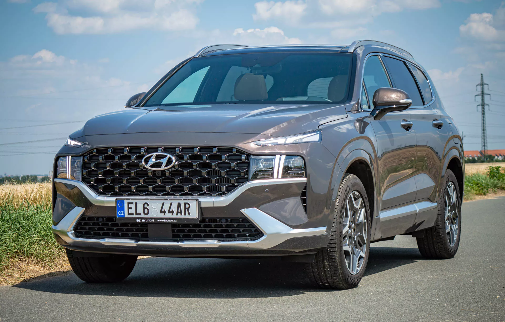 Obrázok zobrazuje SUV Hyundai Santa Fe 2020 zaparkované na asfaltovej ceste, fotografované z ľavého predného trojštvrťového uhla. Výrazne zobrazuje veľkú masku chladiča s charakteristickým vzorom a centrálnym logom, elegantné LED svetlomety a tvarovanú kapotu. Široké čelné sklo ponúka dostatočnú viditeľnosť a bočný profil odhaľuje viaclúčové zliatinové kolesá a spätné zrkadlo na strane vodiča s potenciálne integrovanými smerovkami. Tmavo zafarbené vozidlo vyžaruje sofistikovaný vzhľad s modernými dizajnovými prvkami, vrátane možných predných senzorov alebo kamier.