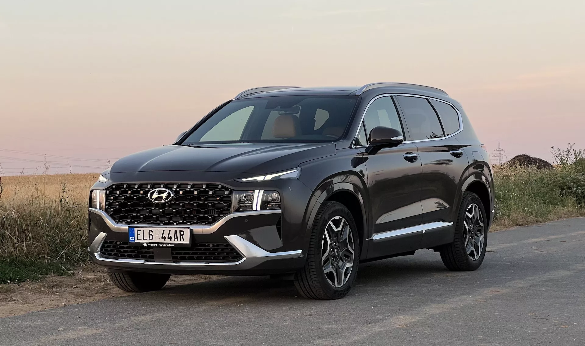 Obrázok SUV Hyundai Santa Fe 2020 zaparkovaného na asfaltovom povrchu, fotografovaný z nízkeho uhla z trojštvrťového pohľadu spredu, zobrazujúci prednú masku, úzke LED svetlomety, nárazník s hmlovými svetlami a falošnou ochrannou doskou, kapotu s ostrými líniami, veľké viaclúčové zliatinové kolesá, čierne ochranné lemovanie blatníkov a spodnej časti dverí, bočné zrkadlo s integrovanými smerovkami a chrómovo lemované okná. Pozadie tvorí neurčitú prírodnú krajinu pod otvoreným nebom.