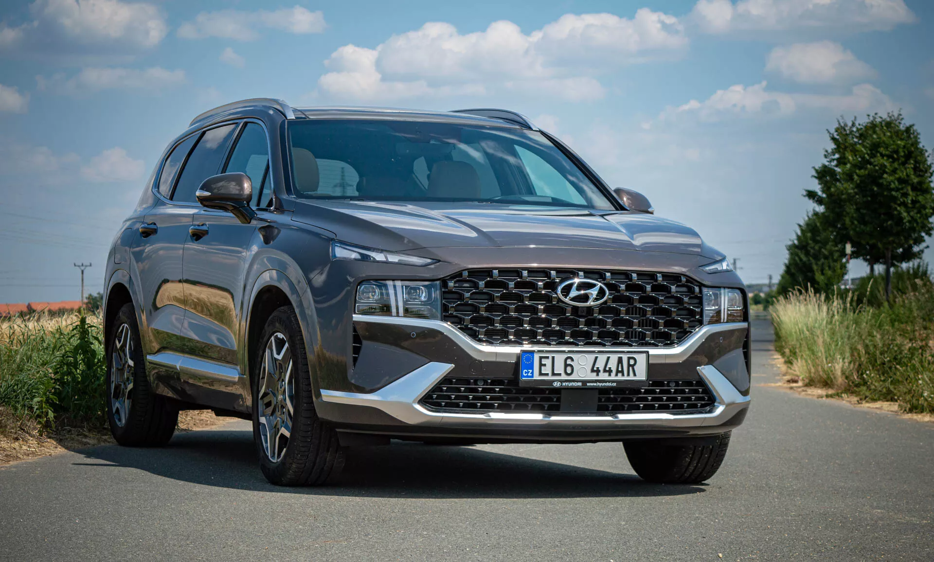 Obrázok SUV Hyundai Santa-Fe 2020 zaparkovaného na dláždenej ceste pod jasnou oblohou s vegetáciou v pozadí. Fotografia je zachytená z prednej trojštvrťovej perspektívy, ukazujúca prednú časť vozidla a ľavý bočný profil. SUV má veľkú mriežku s prepletaným vzorom s logom Hyundai, elegantné uhlové LED alebo HID svetlomety, viacramenné hliníkové kolesá, moderné a aerodynamické telo s hladkými líniami, bočné zrkadlá s integrovanými smerovými svetlami, zatmavené okná, tmavošedú farbu, prednú registračnú značku a vyzerá, že je v perfektnom stave, čo naznačuje, že je buď nové, alebo dobre udržiavané.