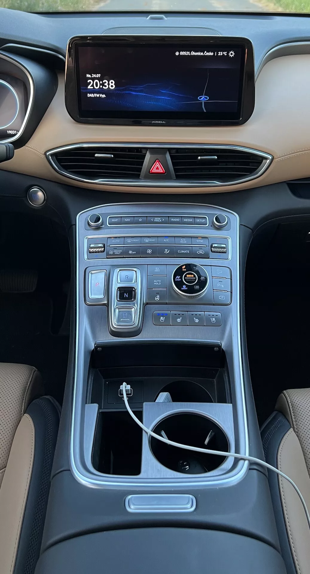 Pohľad zhora na interiér stredovej konzoly Hyundai Santa Fe 2020 zo zadných sedadiel. Obrázok ukazuje modernú palubnú dosku s nočnou cestou zobrazenou na infotainmentovom displeji a digitálnymi hodinami ukazujúcimi '20:38.' Nižšie sú vidieť štíhle horizontálne vetracie otvory, ktoré sa plynule integrujú do palubnej dosky. Stredová konzola obsahuje rad dotykových a digitálnych ovládačov pre klimatizačné a multimediálne funkcie. Lesklý otočný ovládač a susedný prepínače naznačujú pokročilú navigáciu a jazdné režimy. Nižšie je otvorený úložný priestor s viditeľným bielym káblom, čo naznačuje pripojenie zariadení. Stredový tunel, lemovaný kovovým lemom, zvyšuje luxusný dizajn interiéru.